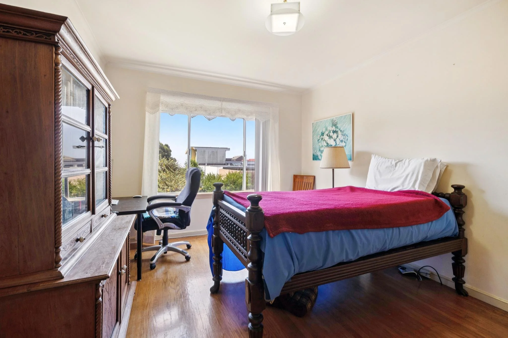 A bedroom with a wooden dresser, an office chair, a bed with a red blanket, a table lamp, a framed floral painting, and a large window with sheer curtains showing trees and neighboring houses outside.