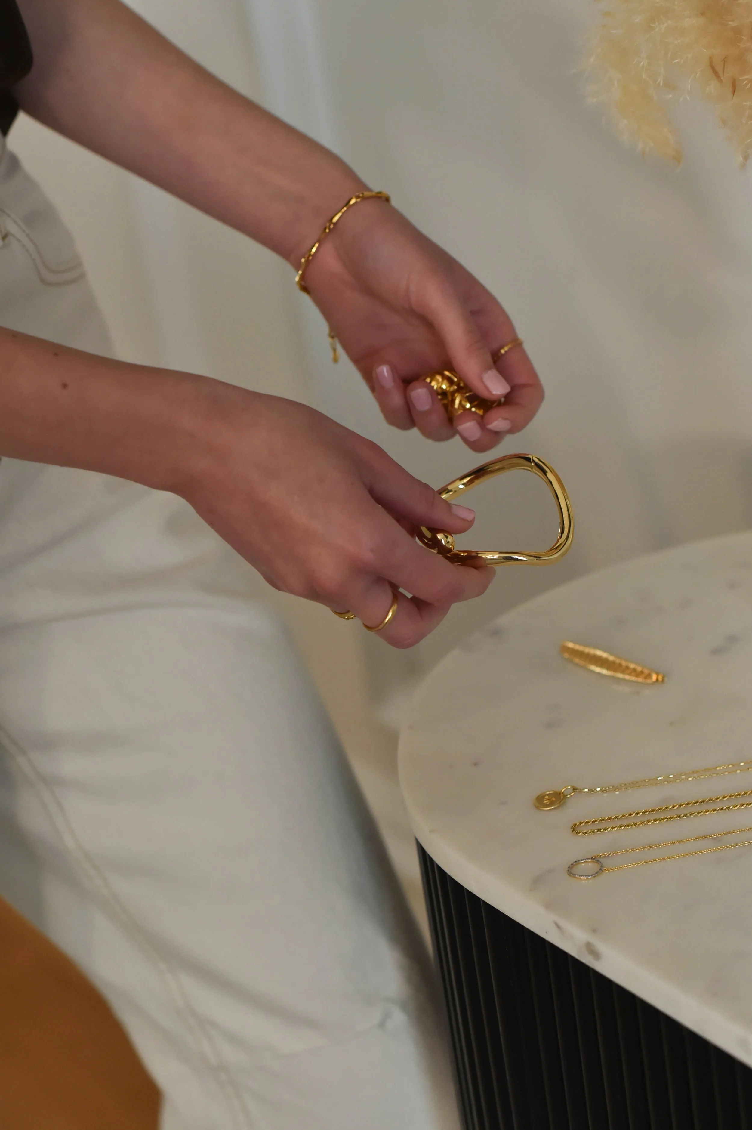 Close-up of a person’s hands and wrists wearing gold jewelry, including rings, a bracelet, and a necklace, with jewelry displayed on a white surface with gold chains.