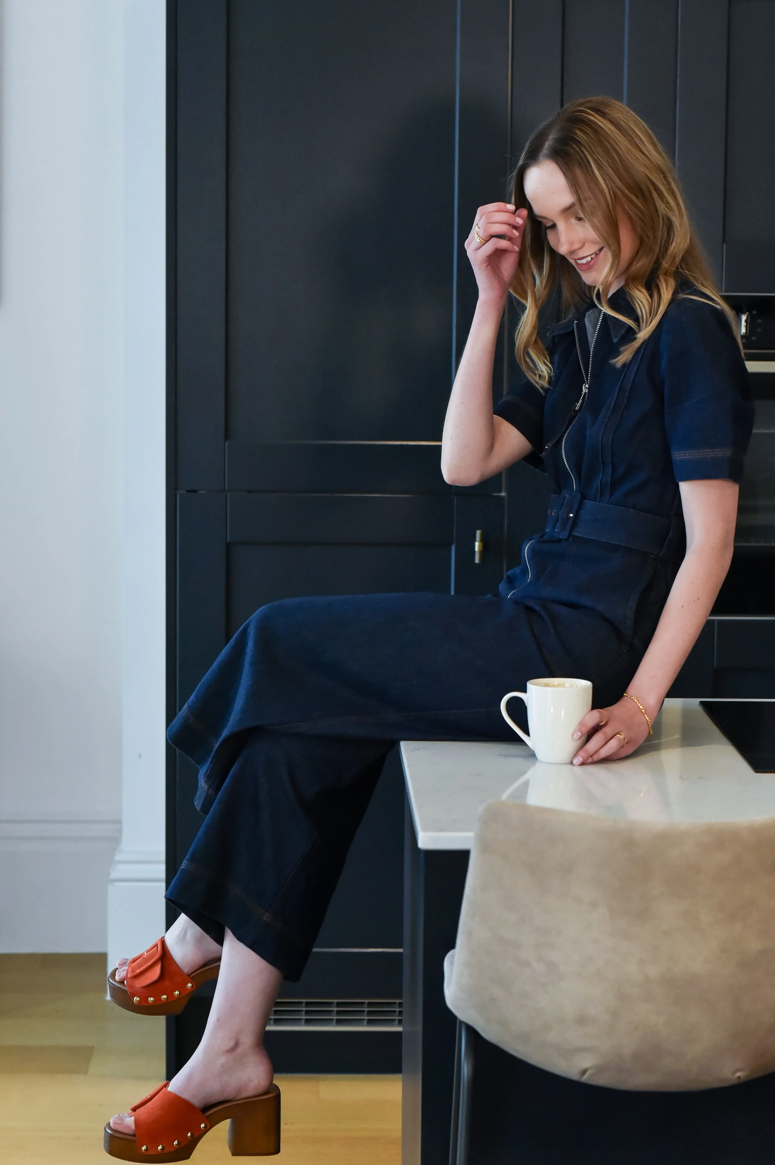 A woman with shoulder-length wavy hair, wearing a dark zip-up dress with short sleeves, is sitting on a kitchen counter holding a white coffee mug. She is smiling and looking down, with one hand touching her hair. She is wearing orange platform sandals with a chunky wooden heel and gold stud details. The kitchen has dark cabinetry and a light-colored marble countertop.