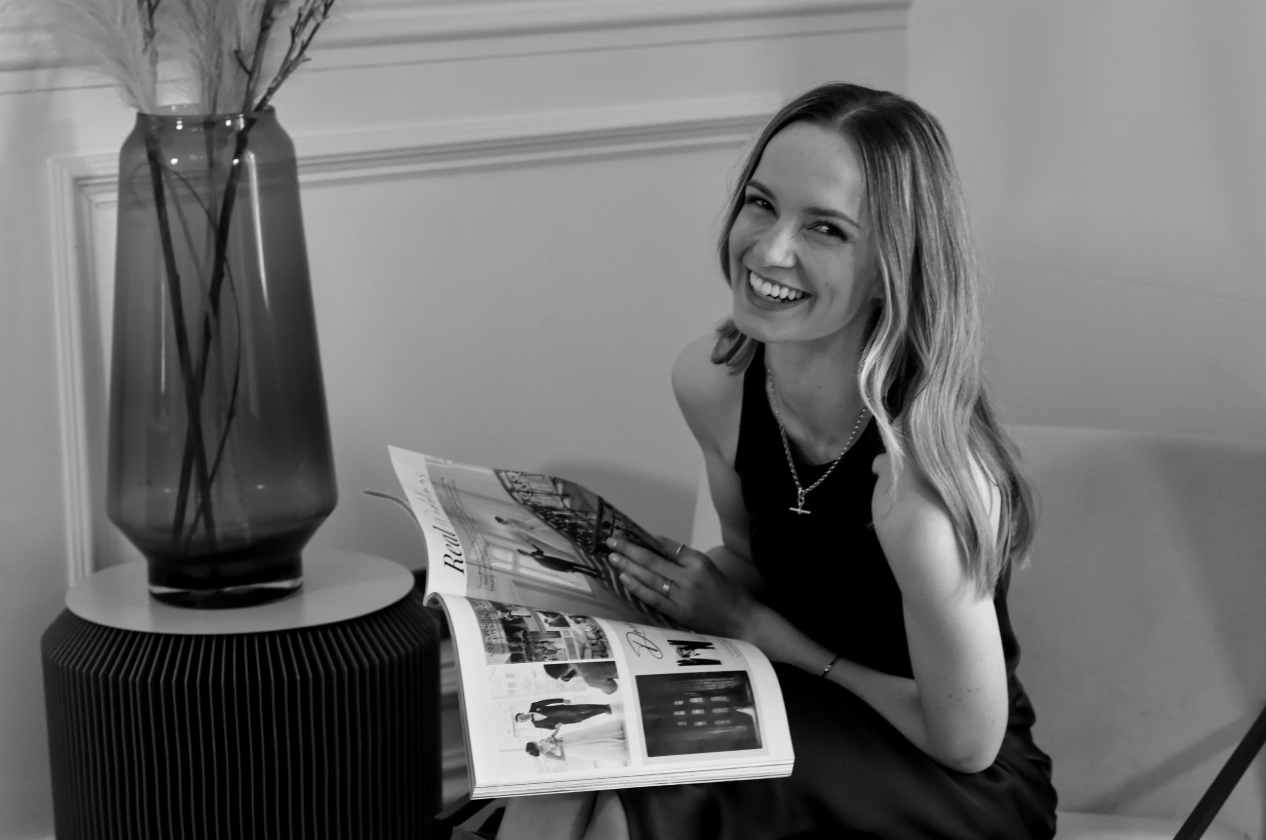 A woman with long, wavy hair smiling, sitting at a table and reading a magazine. There is a tall vase with long-stemmed flowers on the table.