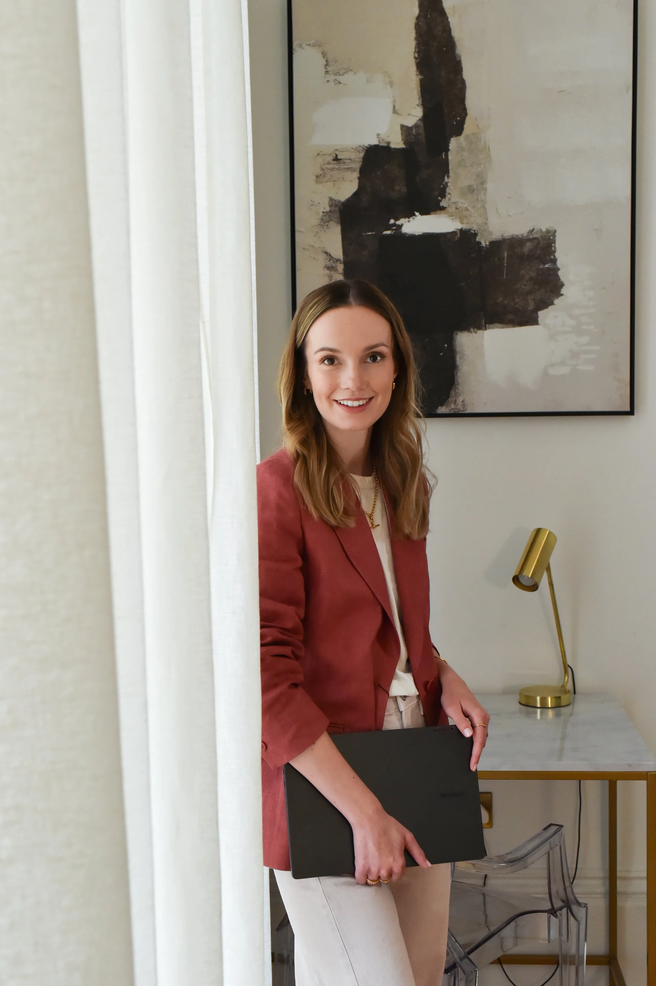 A young woman with shoulder-length light brown hair, wearing a pink blazer and white pants, is holding a closed black laptop and smiling. She is standing in front of a marble countertop in a modern room with abstract artwork on the wall, a gold desk lamp, and transparent chairs.