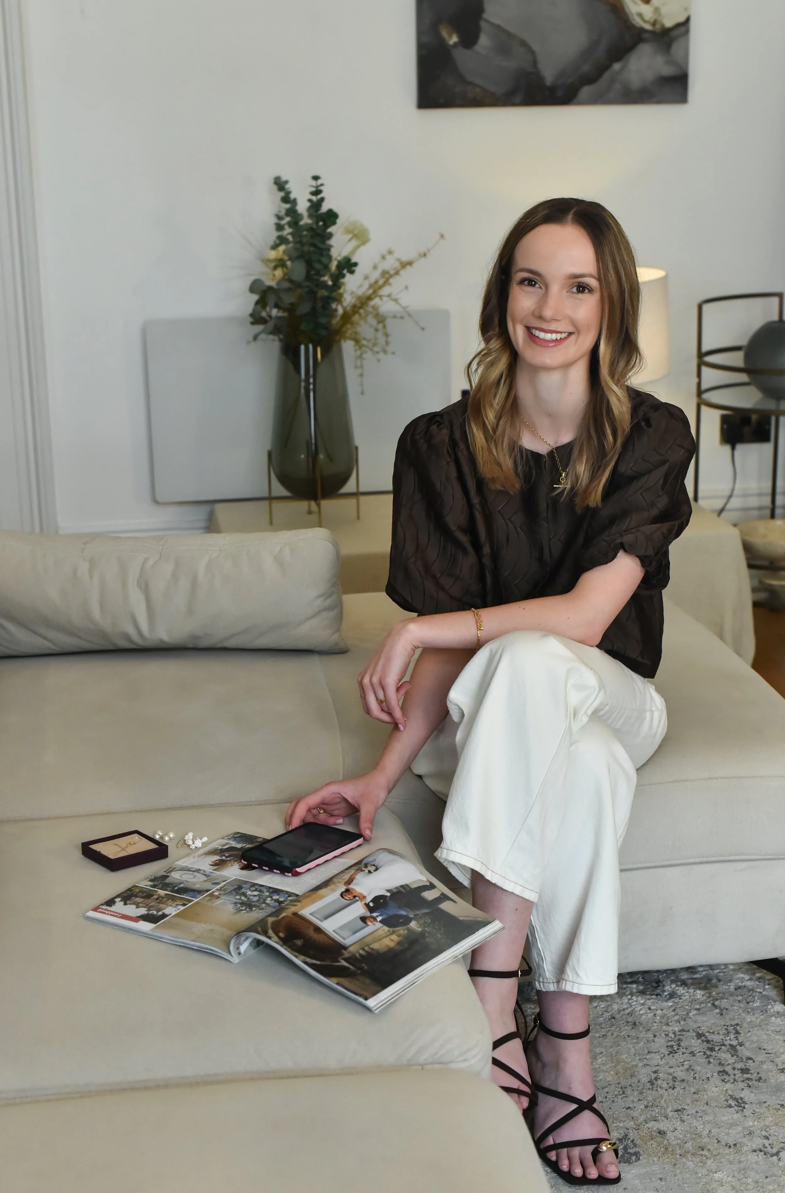 A woman with long brown hair sitting on a beige sofa, smiling at the camera, wearing a dark top and white pants, with black strappy sandals. She is resting her right arm on her knee. In front of her, on the table, are magazines, a smartphone, jewelry, and other small items. The background includes a vase with greenery and flowers, a piece of abstract wall art, and a lamp.