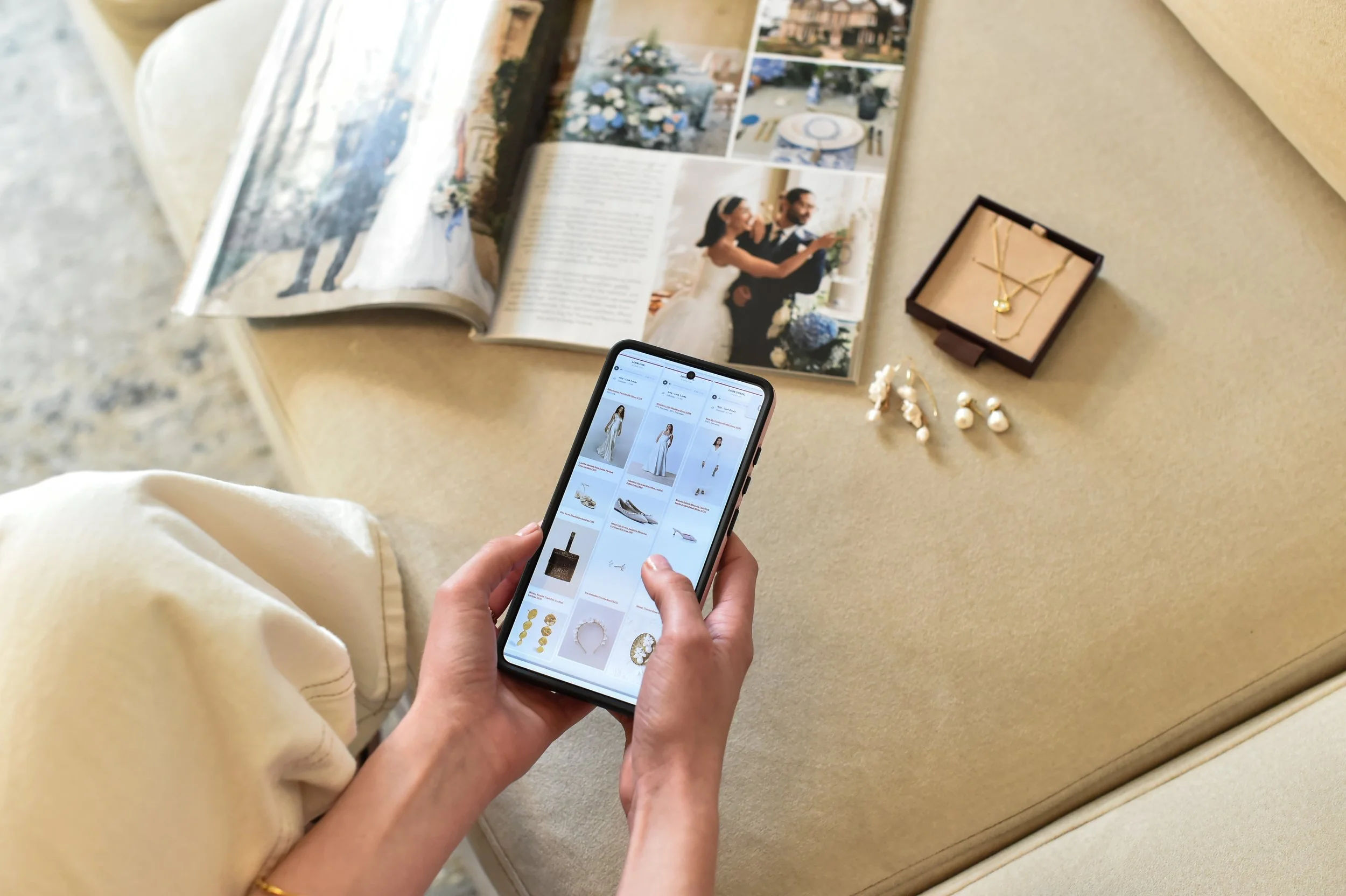 Person browsing jewelry on a smartphone in a living room, with wedding magazine, jewelry box, and jewelry pieces on a coffee table nearby.