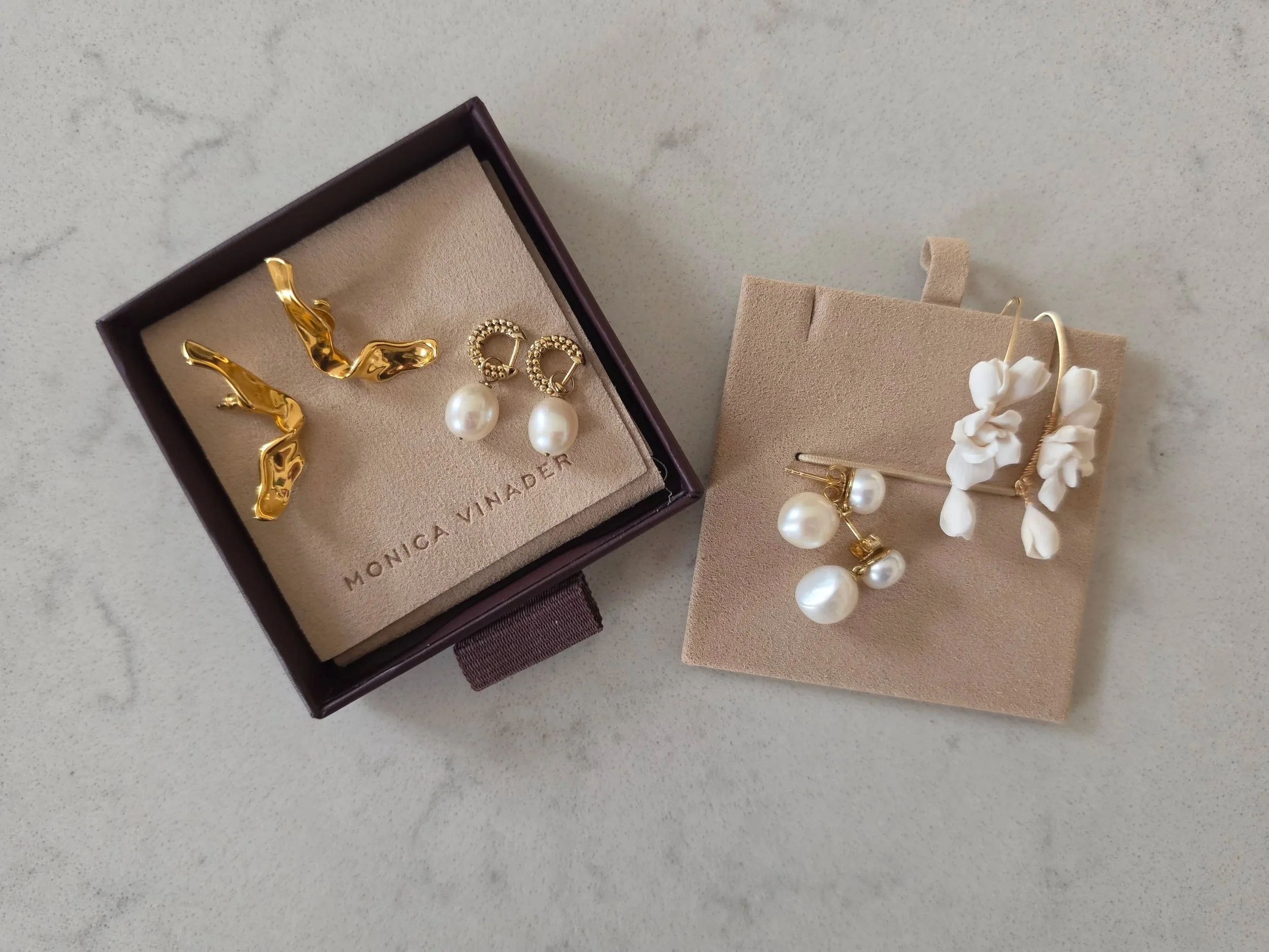 A jewelry box with gold and pearl earrings and a pair of floral earrings beside it on a marble surface.