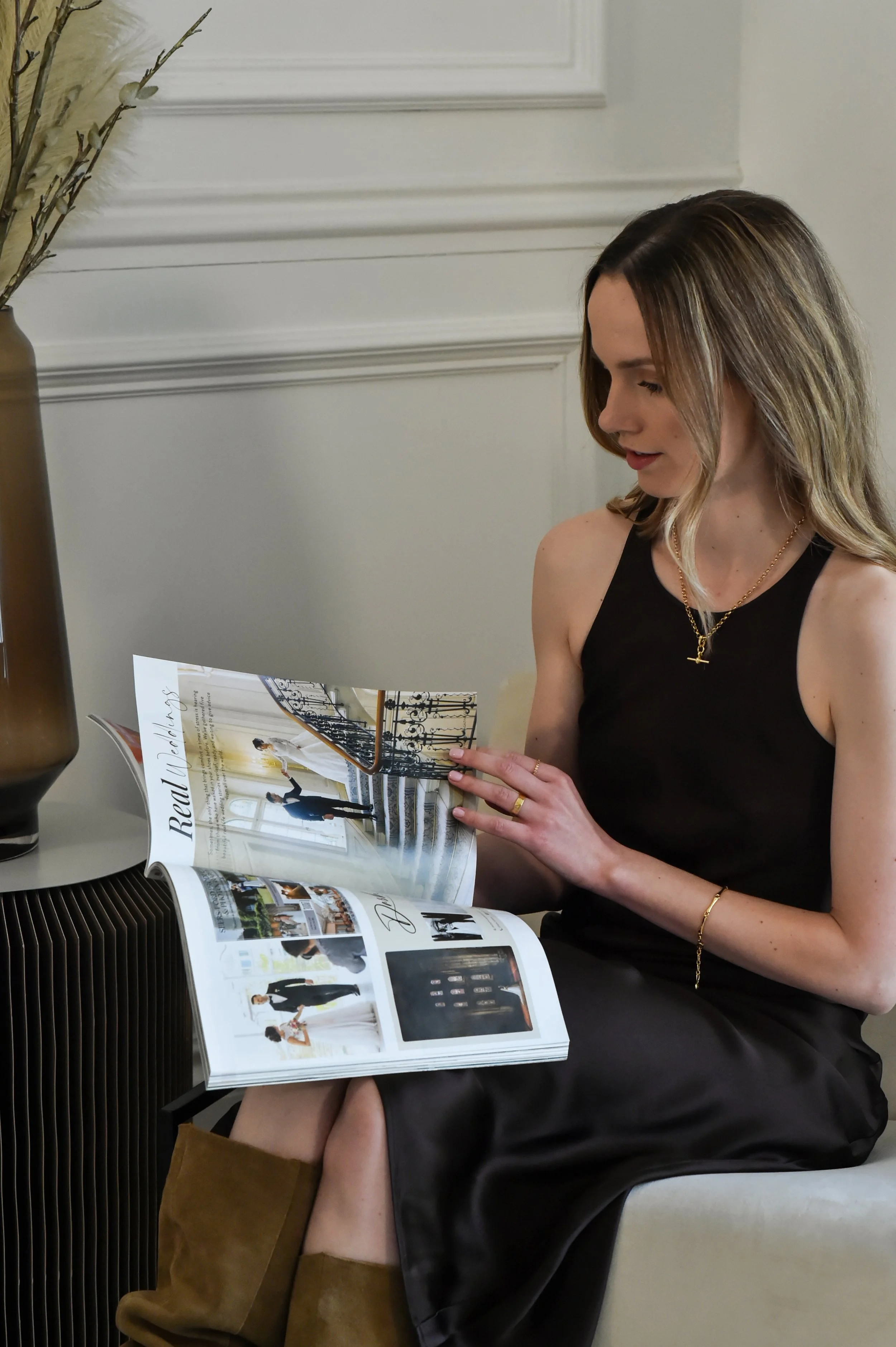 A woman with shoulder-length brown hair reading a magazine. She is sitting on a white chair in a room with gray walls and decorative molding. She is wearing a black sleeveless top, a gold necklace, and brown boots.