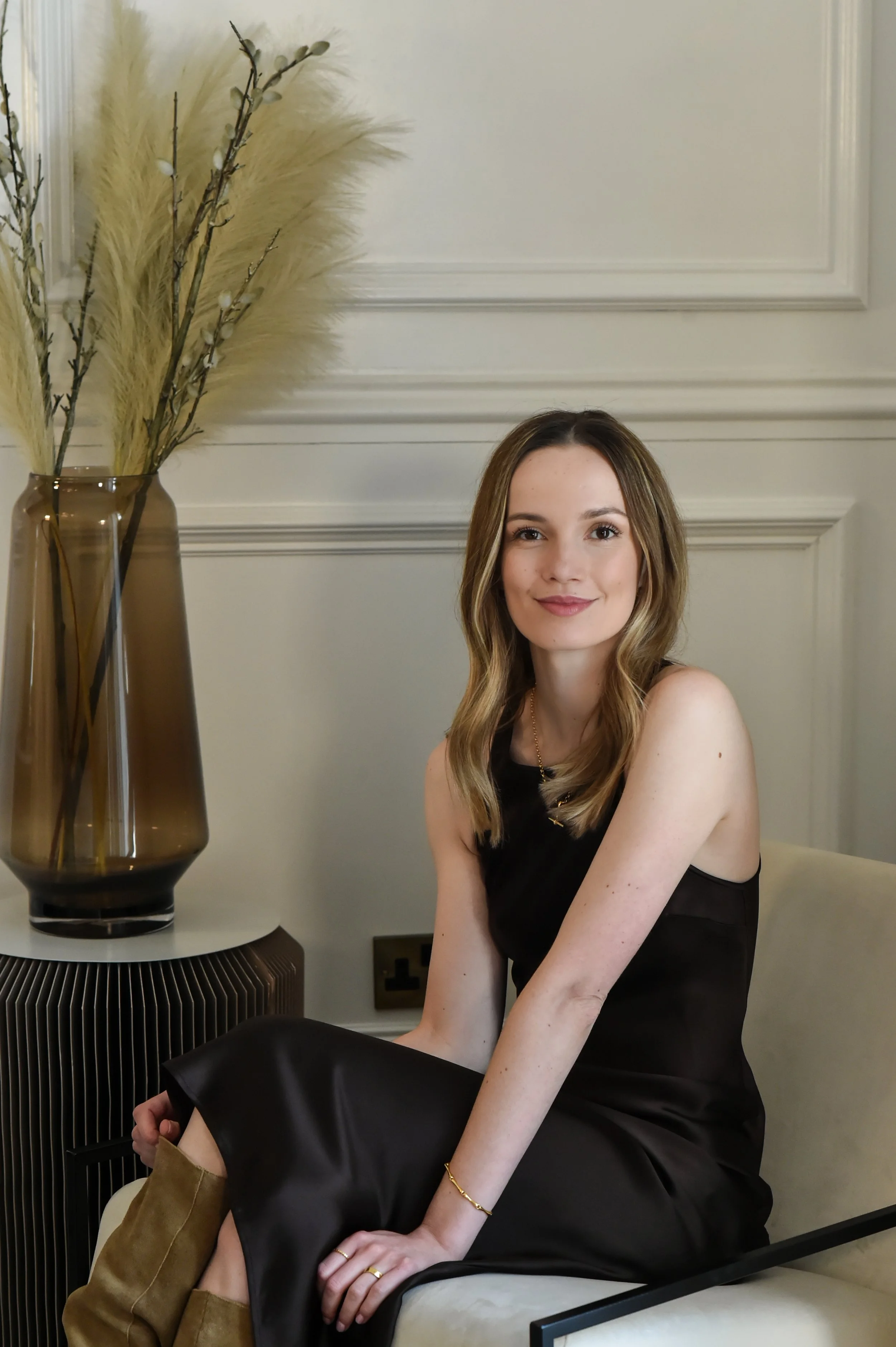A young woman with long wavy brown hair and light skin, sitting on a white sofa in a living room, smiling at the camera. She is wearing a black sleeveless dress and gold jewelry, with some brown boots. There is a tall brown vase with pampas grass and decorative branches on a side table next to her, with a white wall and molding in the background.