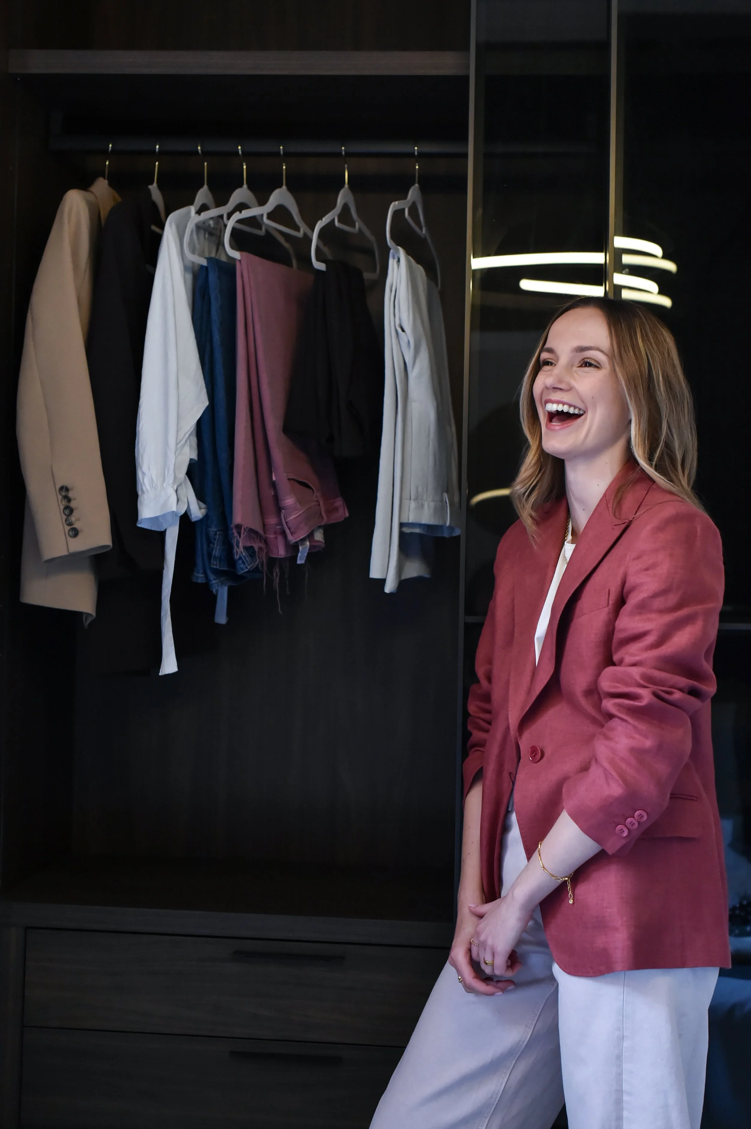 A woman stands in front of an open closet with hanging clothes, smiling and wearing a red blazer, white shirt, and white pants.