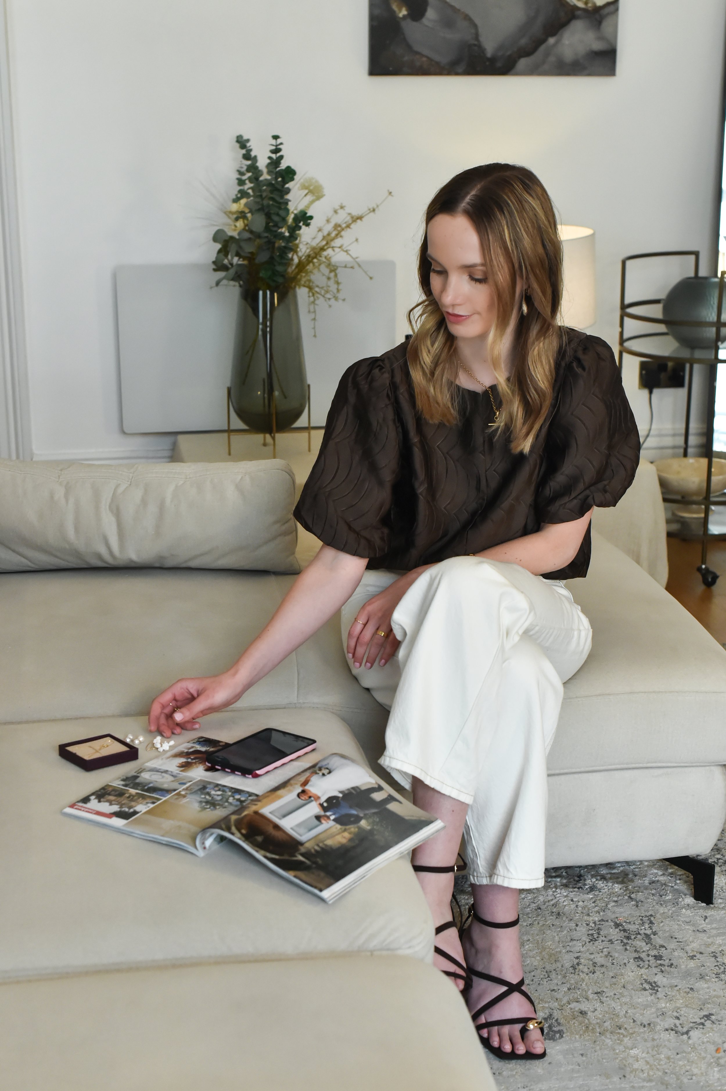 A woman with shoulder-length wavy hair and fair skin is sitting on a beige sofa in a modern living room. She is wearing a black puff-sleeve blouse, white pants, and black strappy sandals. Her head is slightly tilted downward as she looks at a display of jewelry and magazines on the table in front of her. The room features a large vase with greenery, a lamp, and artwork on the wall.