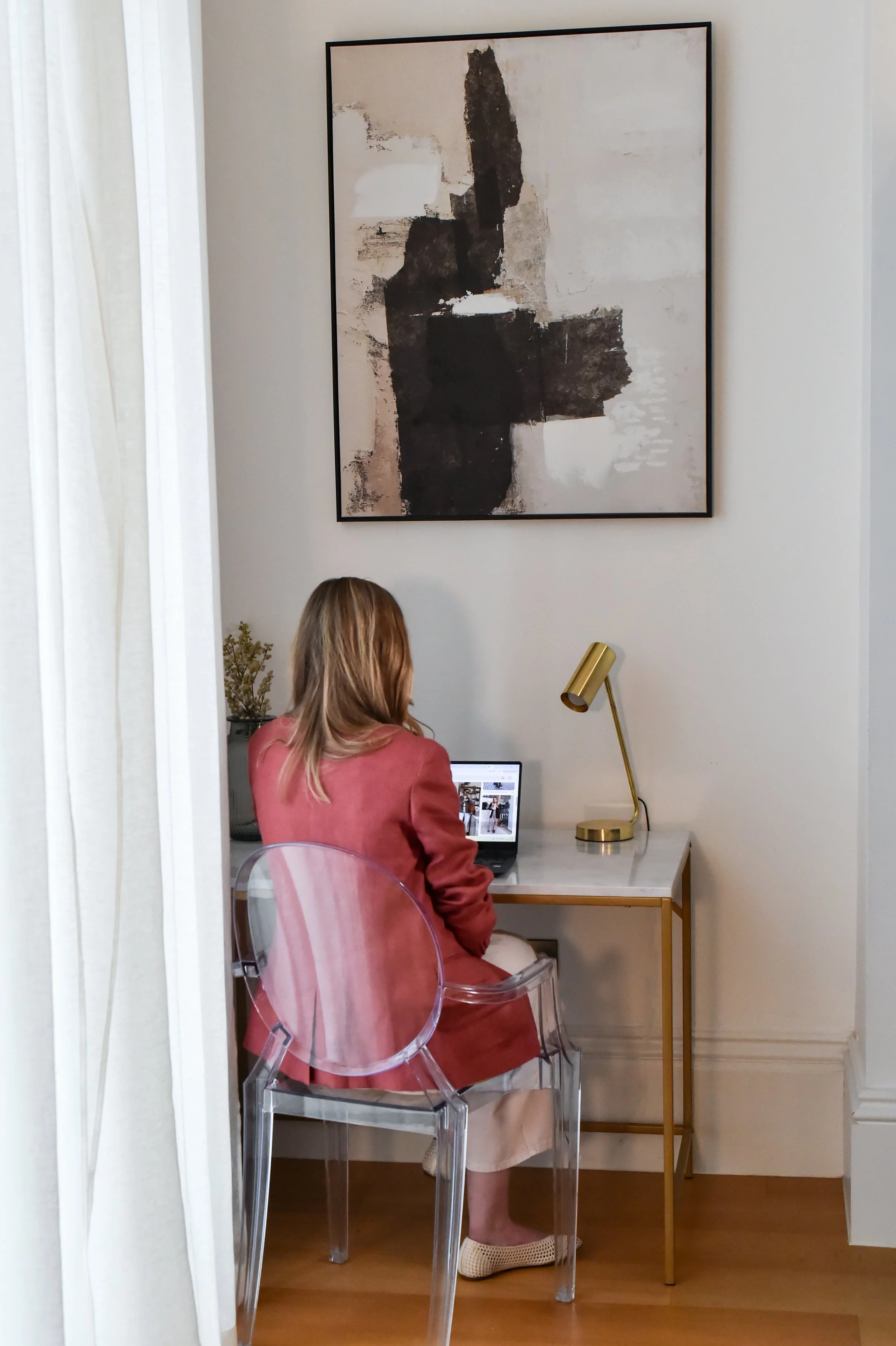 A woman is sitting at a desk with a laptop, working in a room with modern decor. There is an abstract painting on the wall above the desk, and a gold desk lamp next to her. She is dressed in a pink blazer and beige skirt, wearing a beige shoe and transparent chair.