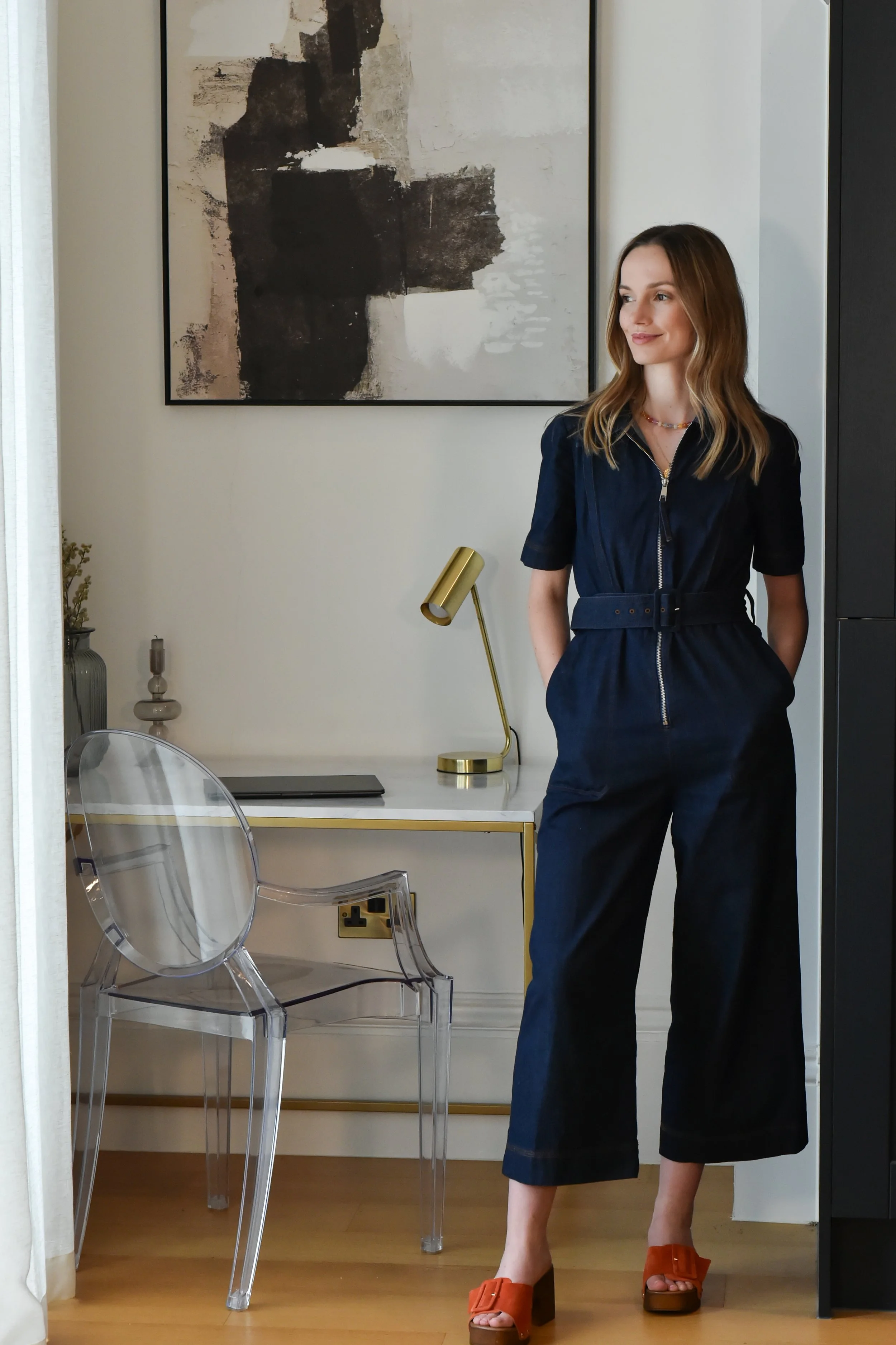 A woman with long, light brown hair standing in a modern, well-lit room. She is wearing a dark blue jumpsuit with a belt and orange high-heeled sandals. Behind her, on the wall, there is an abstract black, white, and beige painting. There is a white desk with a black laptop, a gold desk lamp, and some decorative items on it. Transparent chairs are positioned near the desk, and there is an electrical outlet on the lower wall.