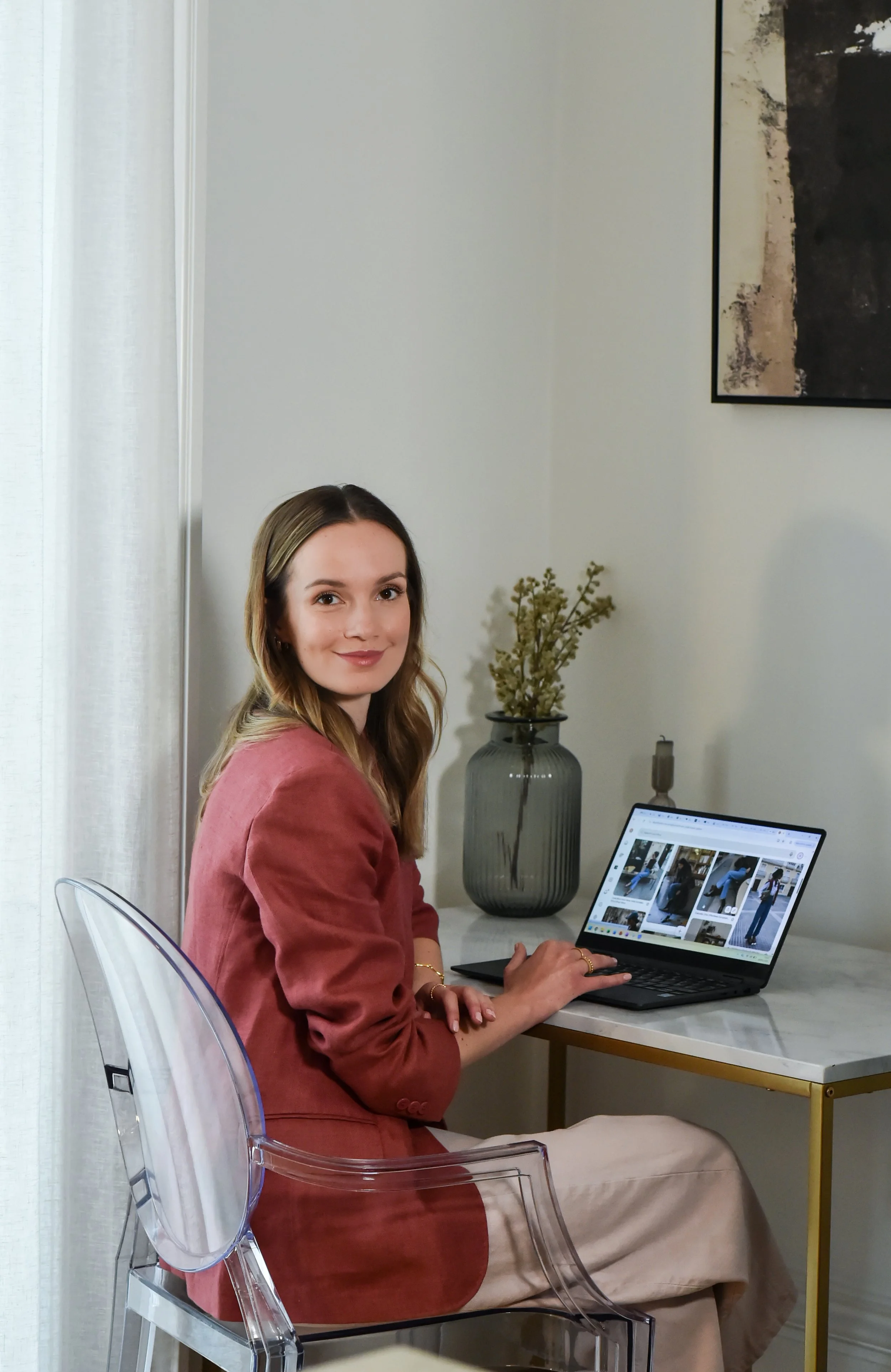 A young woman with light brown hair and a pink blazer sitting at a white desk with a laptop, smiling at the camera in a bright room with white curtains, a large black and white painting on the wall, a gray vase with dried flowers, and a candle.