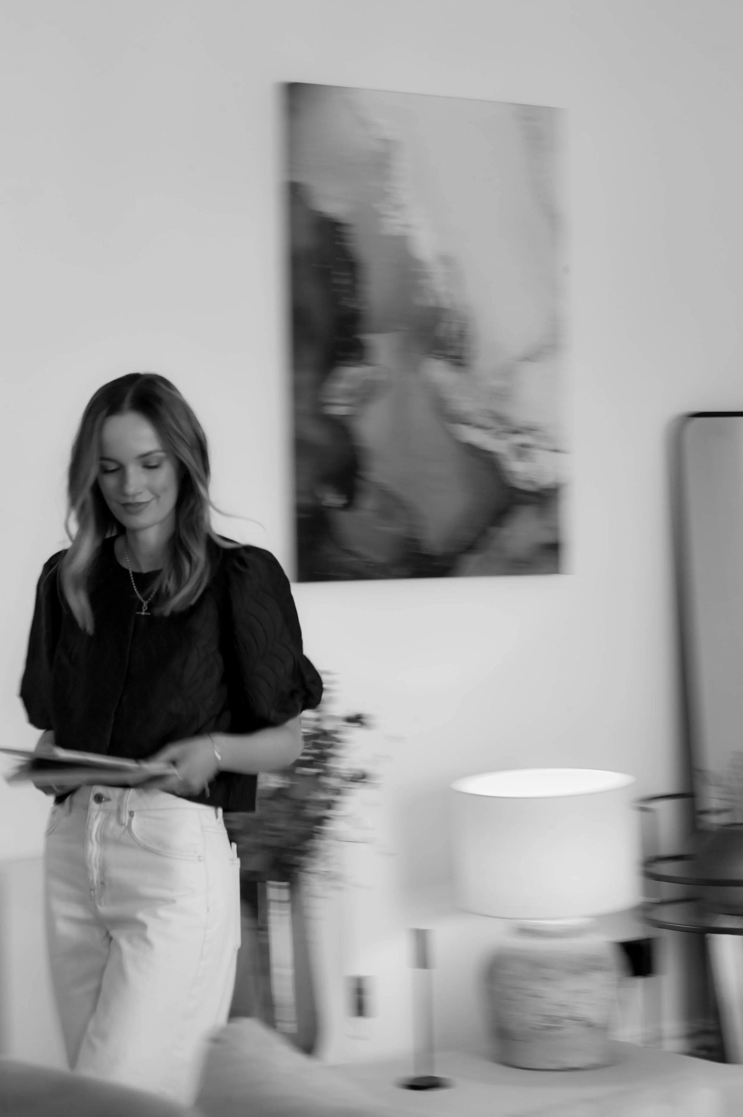 A woman with long wavy hair wearing a black top and white pants standing in a room with white walls, holding a book or magazine, with a blurred artwork on the wall behind her.