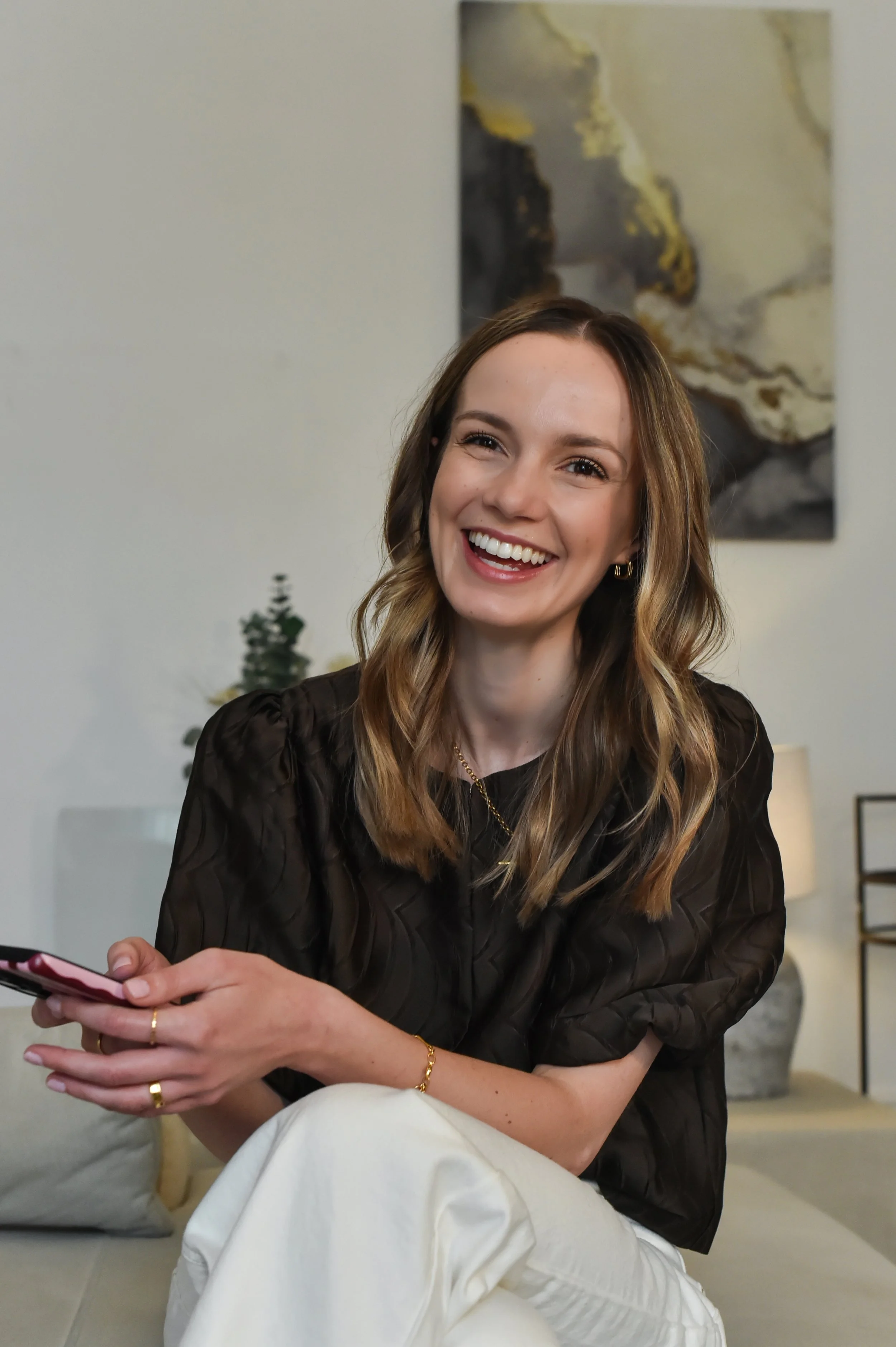 A woman with wavy, shoulder-length brown hair and light skin, smiling while seated, holding a smartphone in her hands. She is wearing a black, textured blouse, gold jewelry, including earrings, a bracelet, and rings. The background includes a small plant, a painting with abstract patterns, and a part of a beige-colored room.