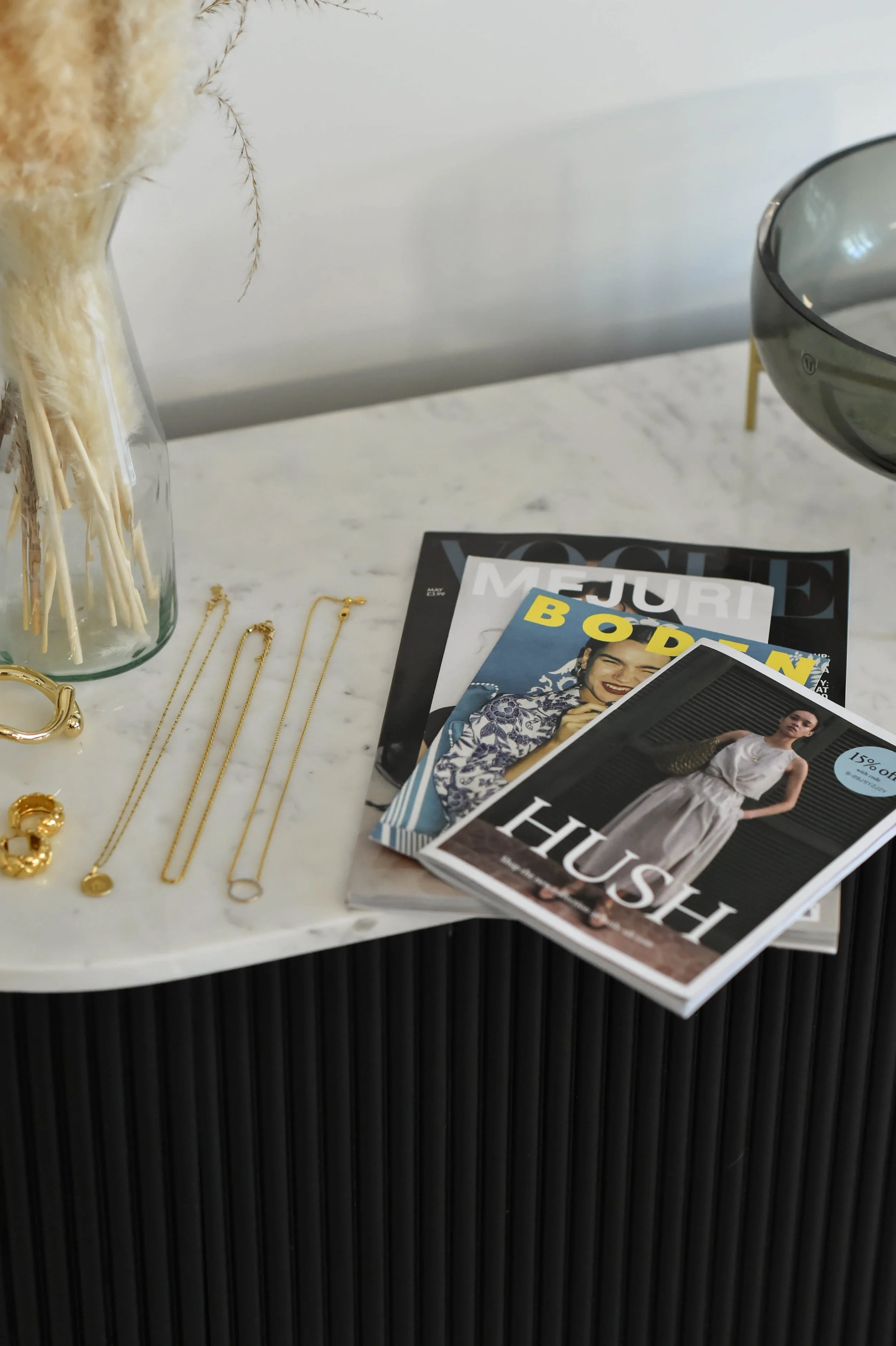 A white marble table with gold jewelry, magazines, and a glass vase with dried plants on top.