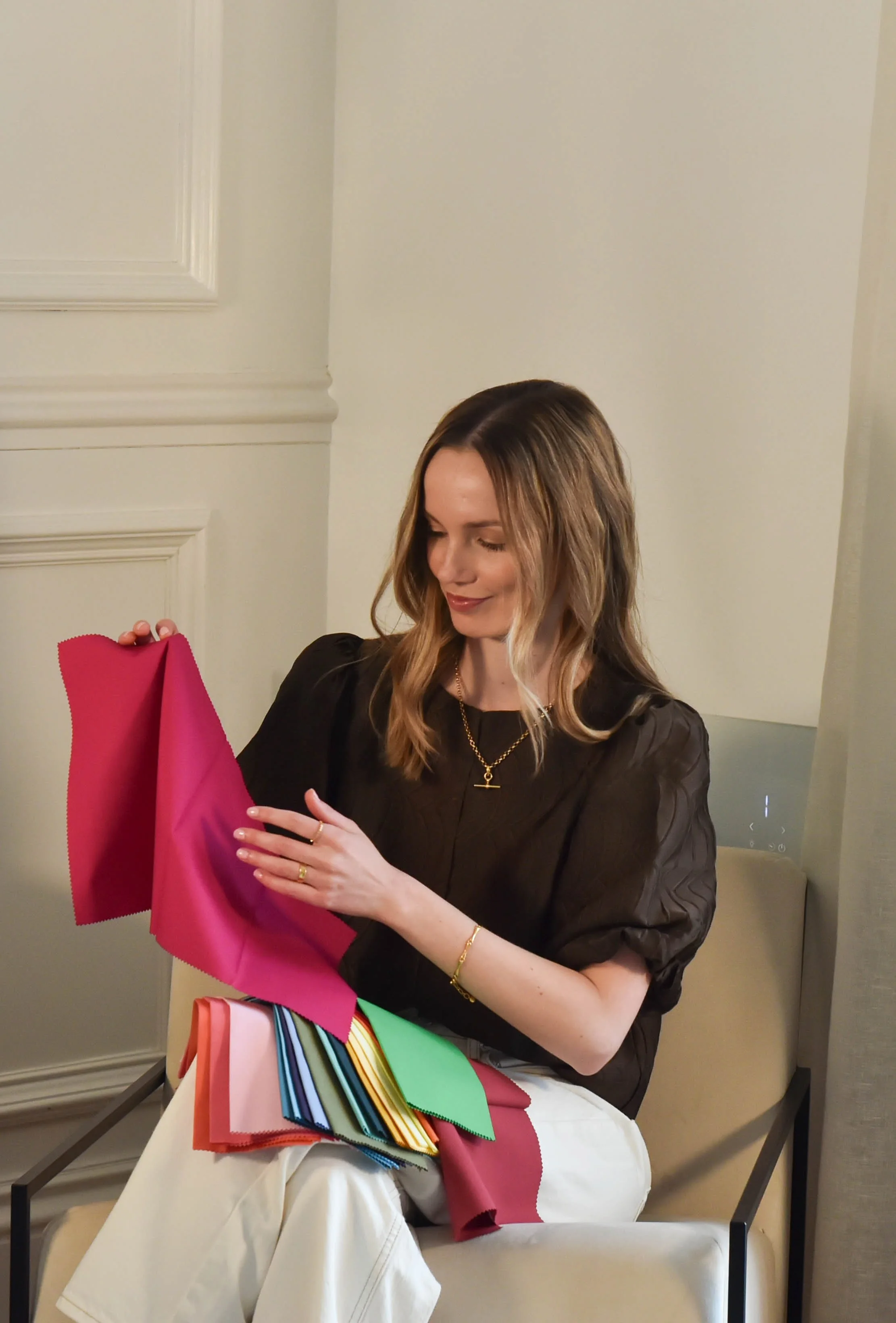 Woman with long hair sitting on a beige chair, holding colorful fabric swatches and a pink piece of fabric, looking down with a smile.