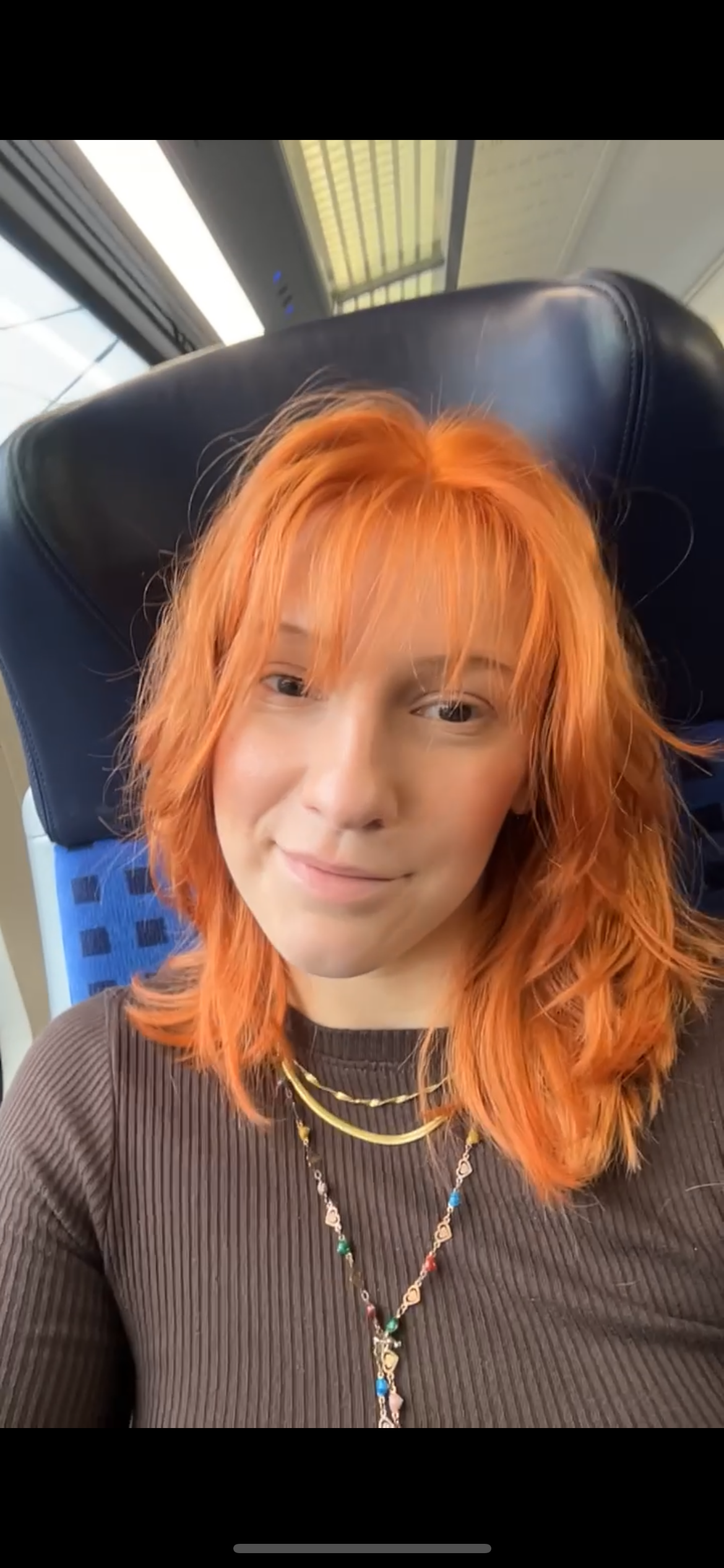 Junge lächelnde Frau mit orangefarbenem, schulterlangem Haar und Pony sitzt in einem Zugsitz und trägt einen dunkelbraunen Pullover und mehrere goldene Halsketten.
