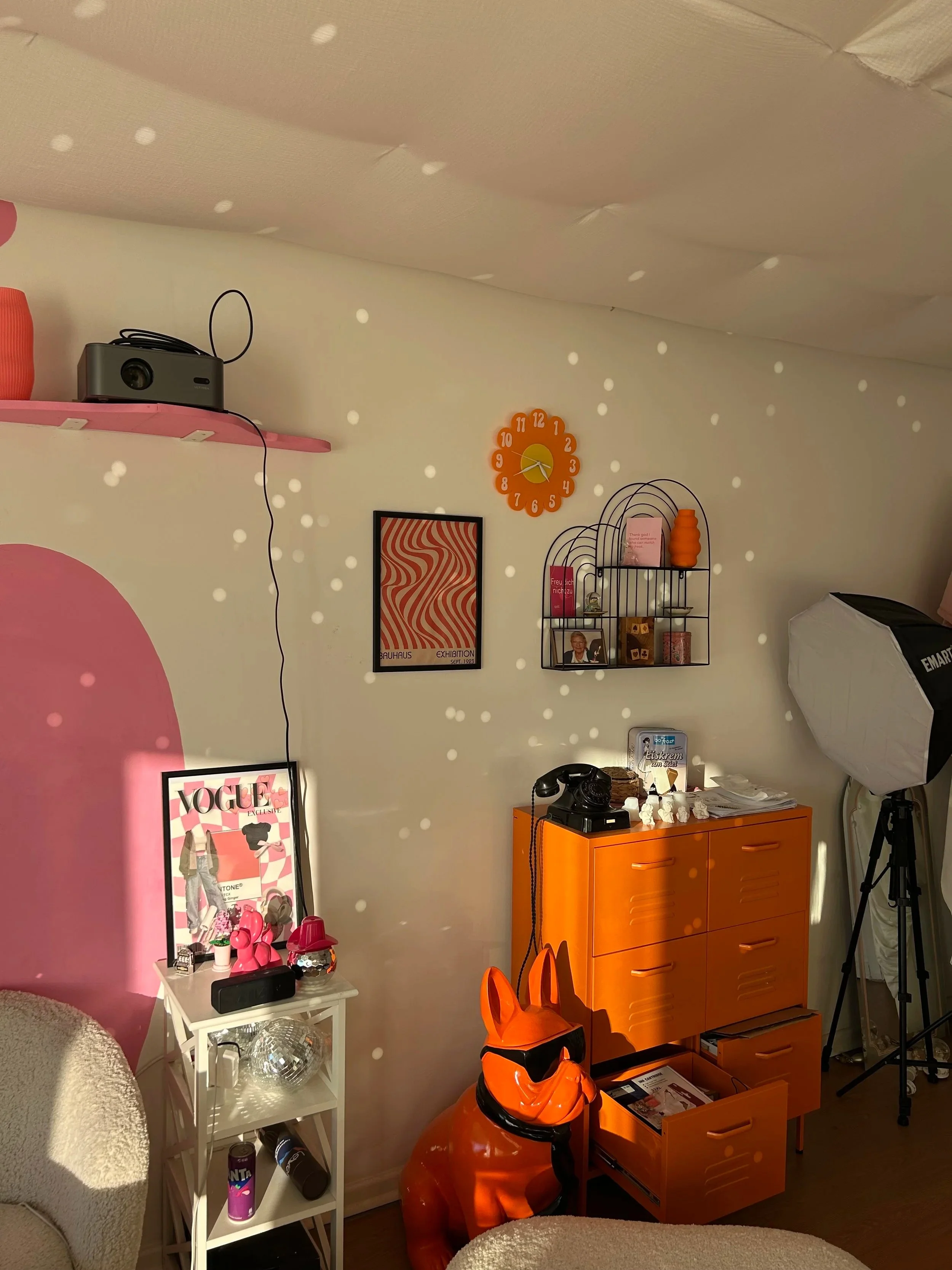 Auschnitt eines Wohnzimmers mit orangener und rosafarbener Deko, welches in warmem Sonnenlicht angestrahlt wird und Diskokugeln helle Lichtreflektionen an die Wand scheinen.