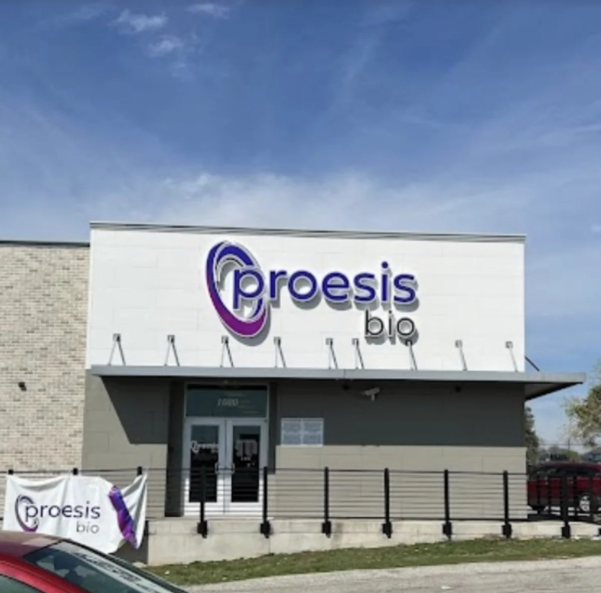 storefront sign for Proesis Bio with a purple and gray logo, glass entrance door, and a small banner in front also displaying the Proesis Bio logo.