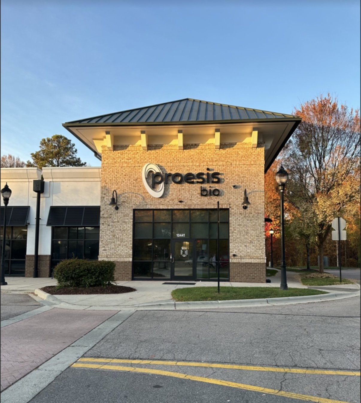 The exterior of a Proesis Bio building with a brick facade, large windows, and a sign with the company's logo and name. The background shows trees with autumn leaves and a clear sky.