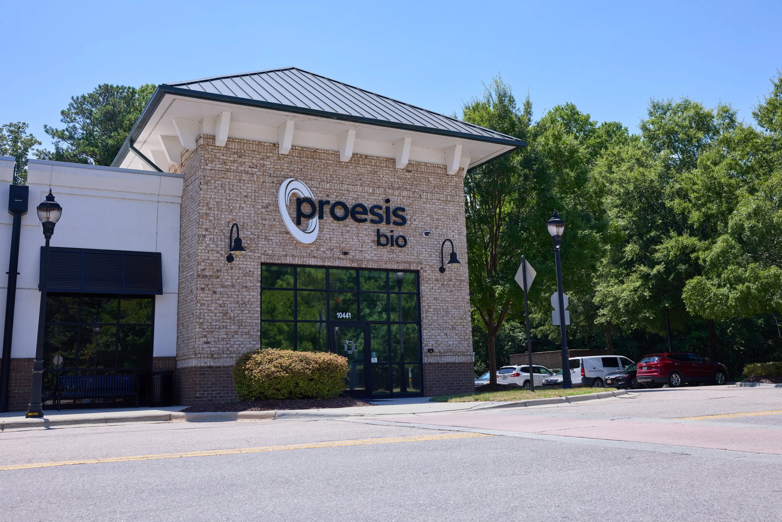 Exterior view of a Proesis Bio storefront with a brick facade, large window, and signage. There are trees and parked cars along the street under a clear blue sky.