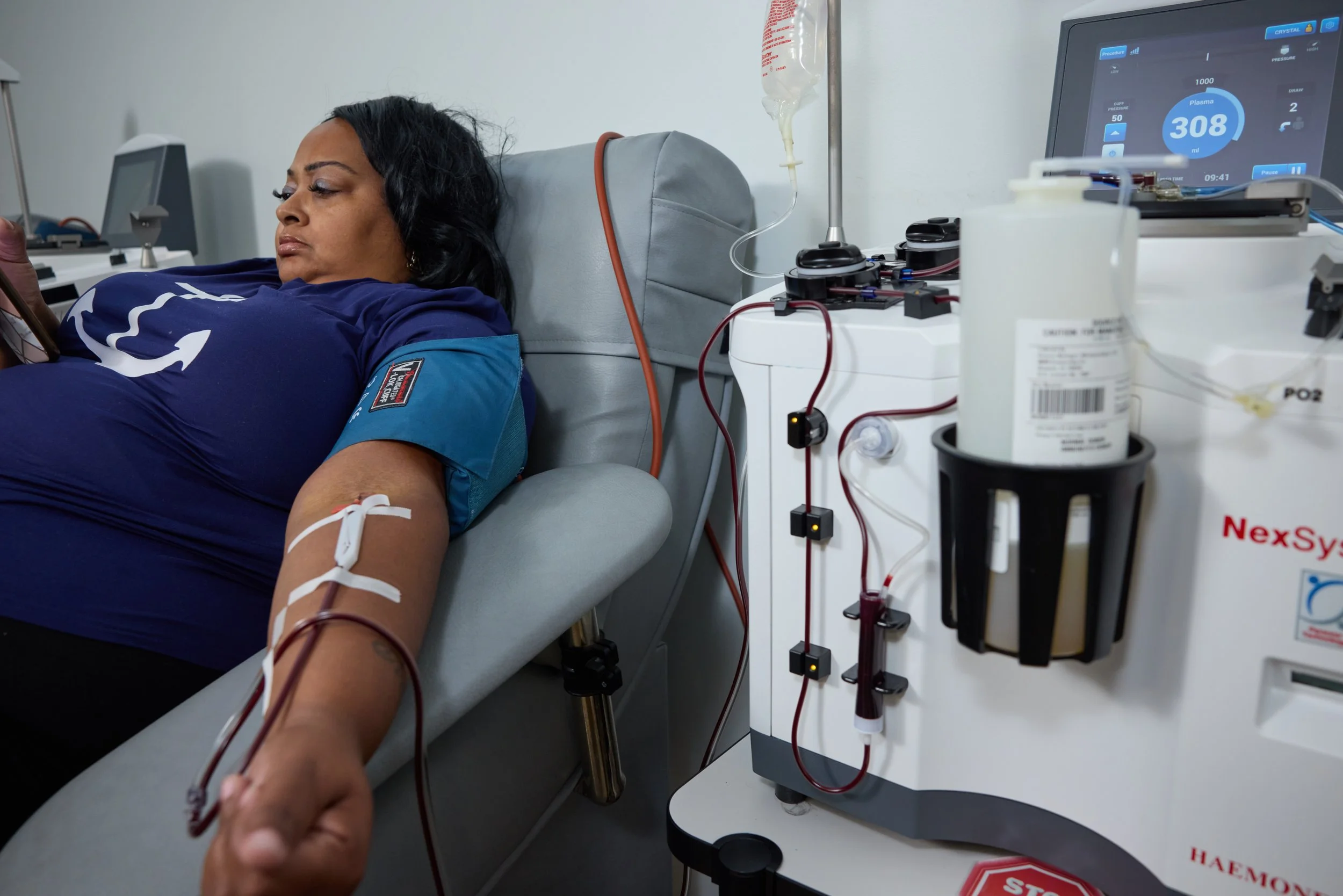A woman in a blue shirt lying on a medical bed donating blood with medical equipment and a monitor displaying blood volume.