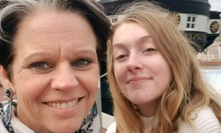 A smiling middle-aged woman with short dark hair and a young woman with long red hair, taking a selfie together outdoors near a boat.