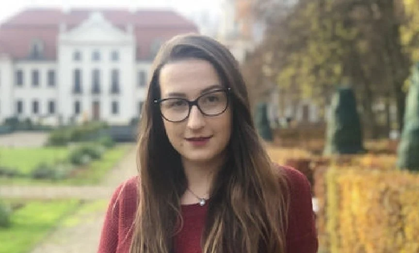Young woman with long brown hair and glasses standing outdoors with trees and a large white building in the background.