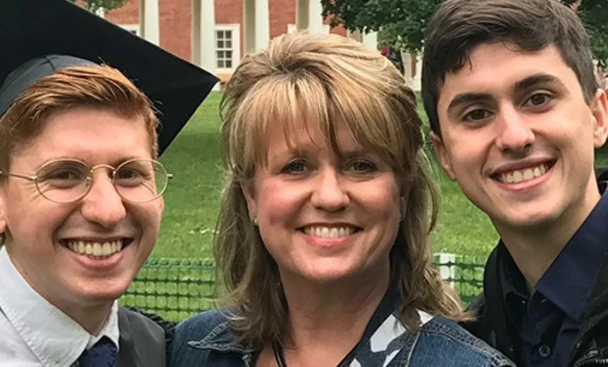 Three people smiling outdoors, one wearing glasses and a graduation cap, in front of a green metal fence with trees and buildings in the background.