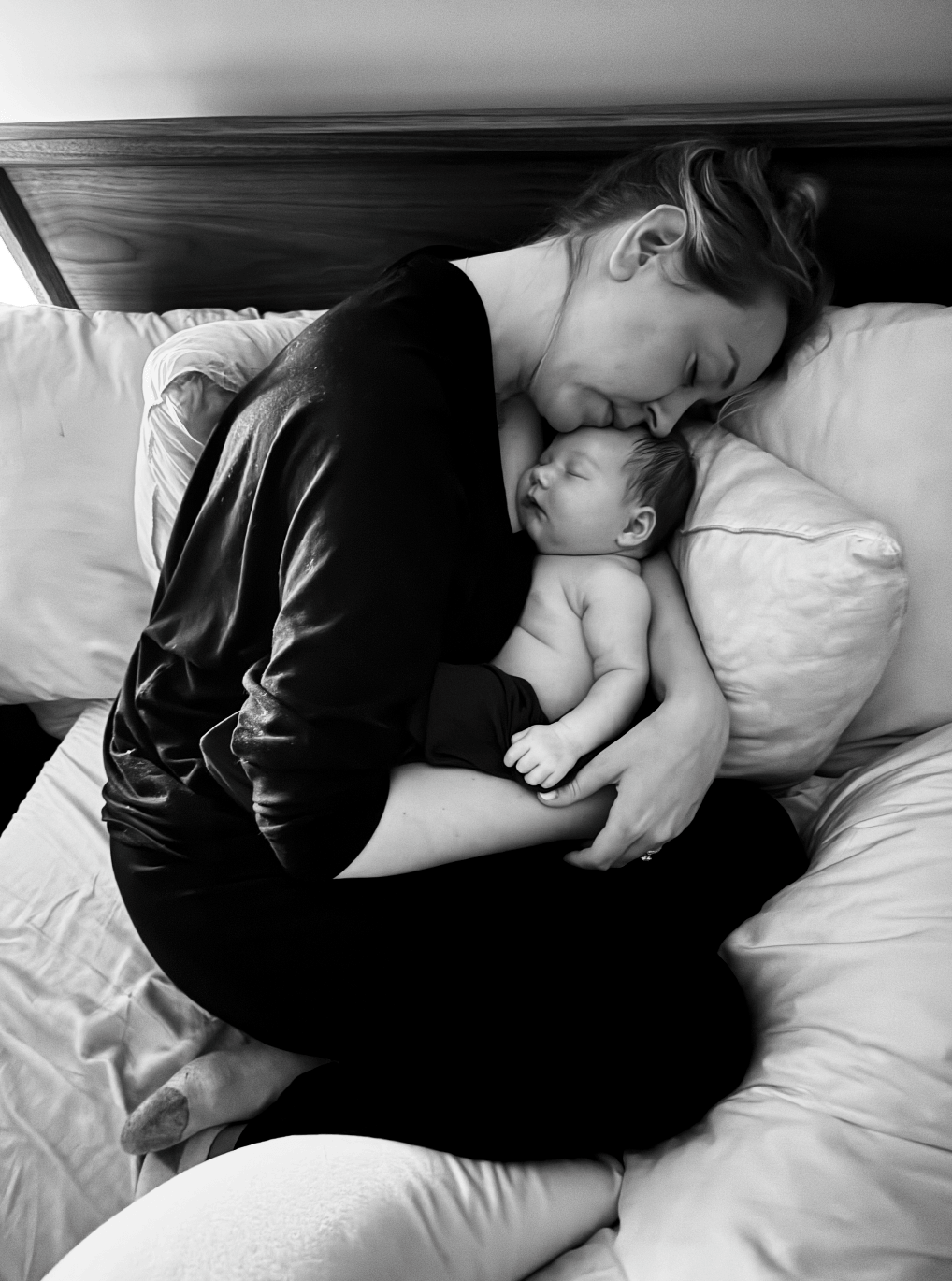 A woman tenderly lying on a bed, holding a sleeping newborn baby close to her chest with her eyes closed and facing each other.