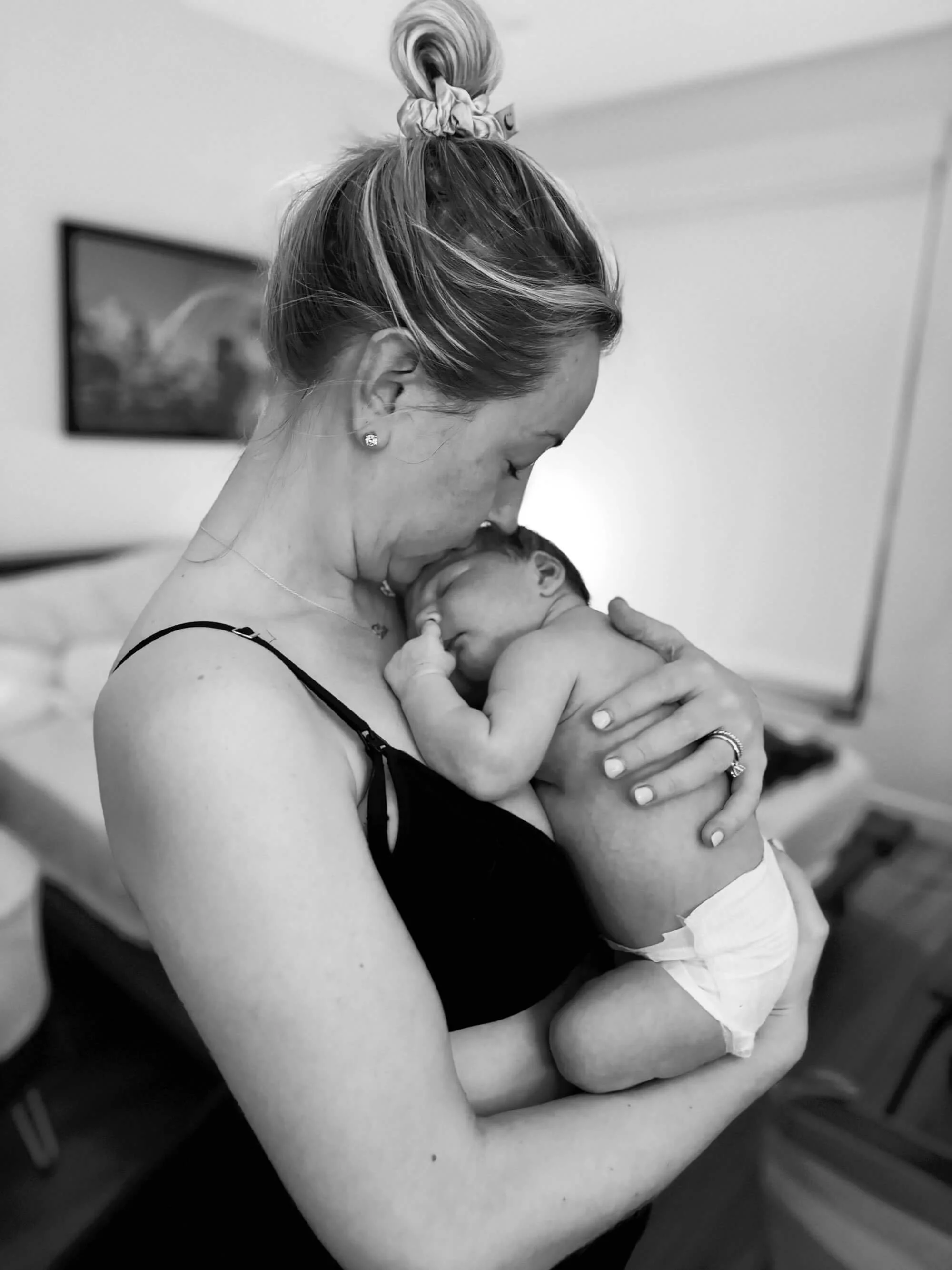 A woman holding a newborn baby close to her chest in a hospital room, the woman is wearing a nightgown and jewelry, the background includes a picture on the wall.