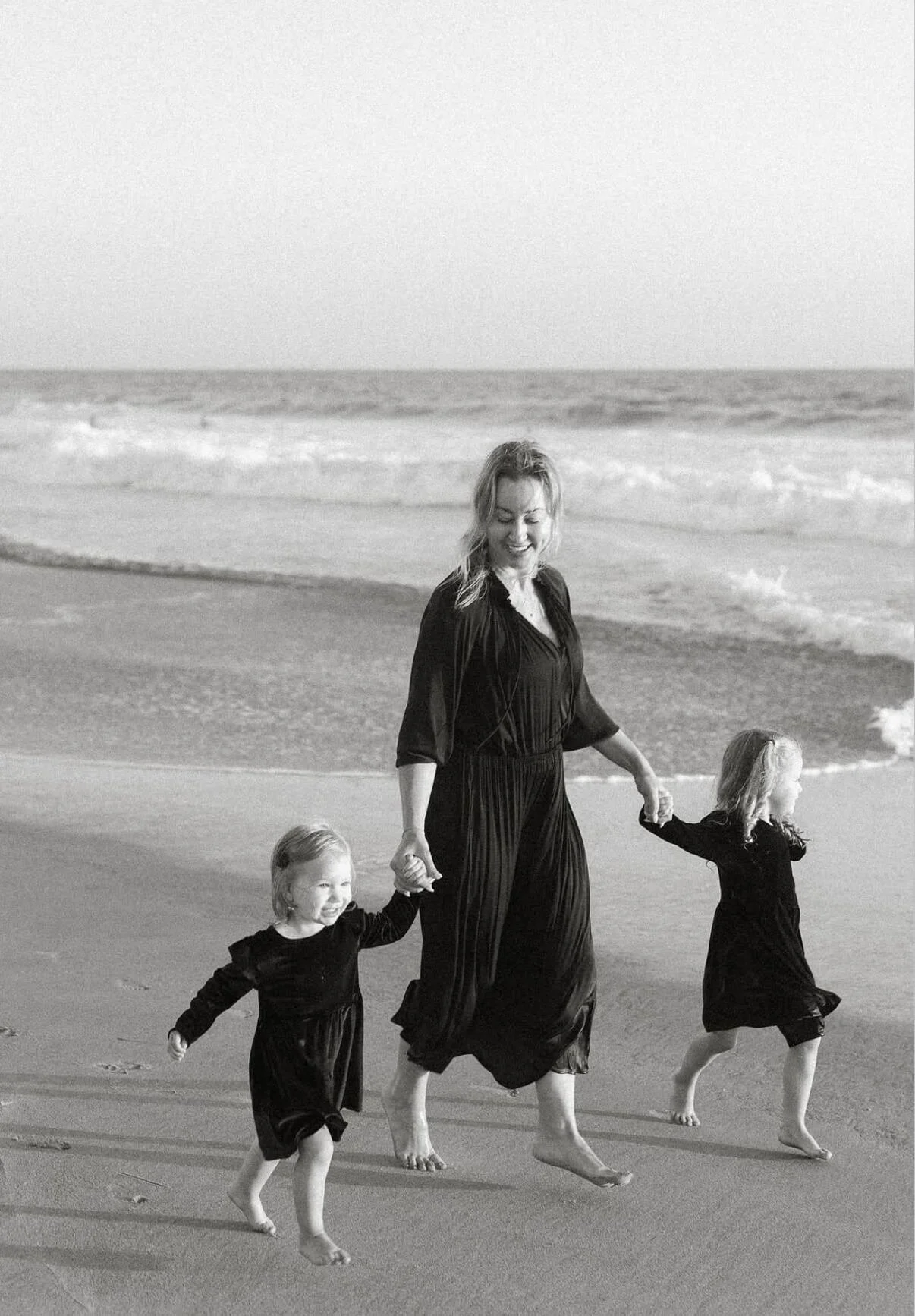 A woman with two young girls walking along the beach at sunset, holding hands and smiling.