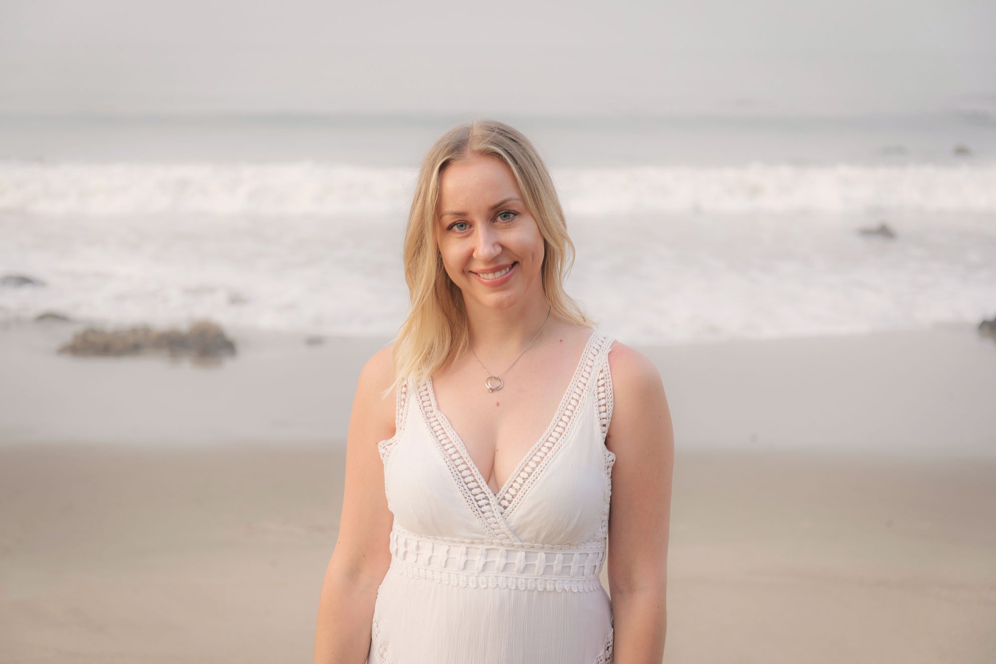 A woman with blonde hair smiling on a beach with the ocean in the background.