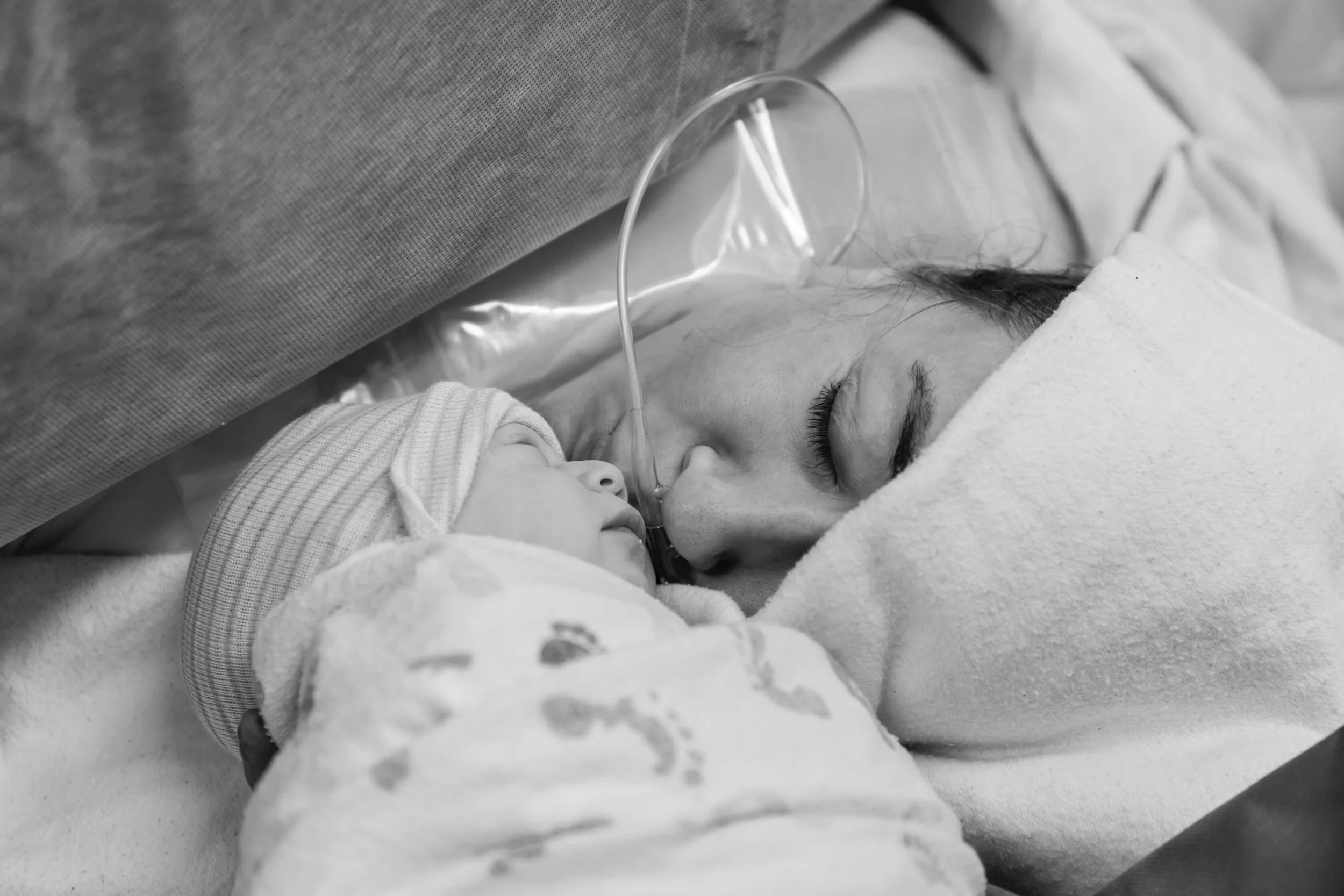 A woman with a nasal oxygen tube lying in bed, resting her forehead against a child's forehead, both sleeping peacefully.