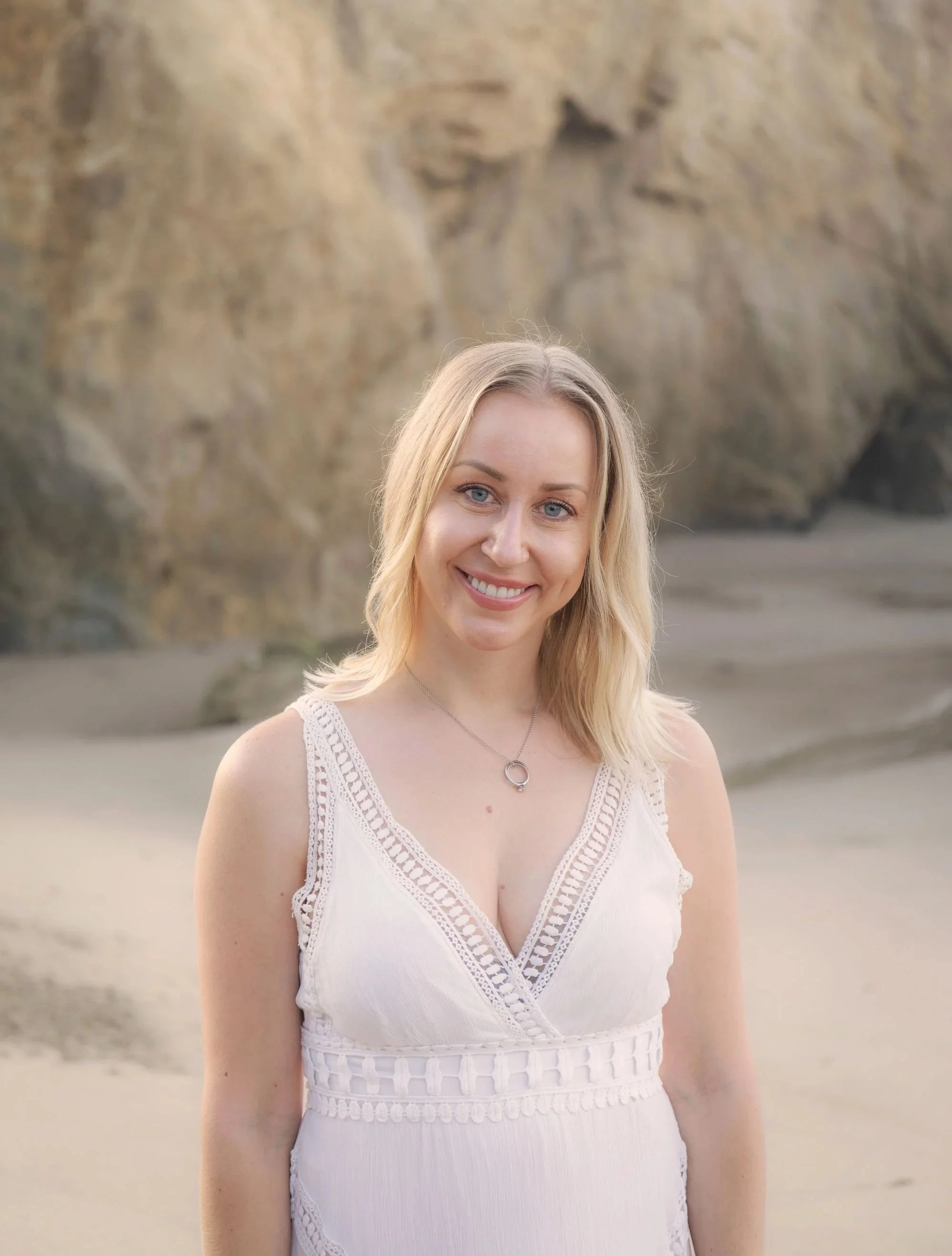 A young woman with blonde hair and blue eyes wearing a white sleeveless dress with lace details, smiling outdoors near rocky formations.