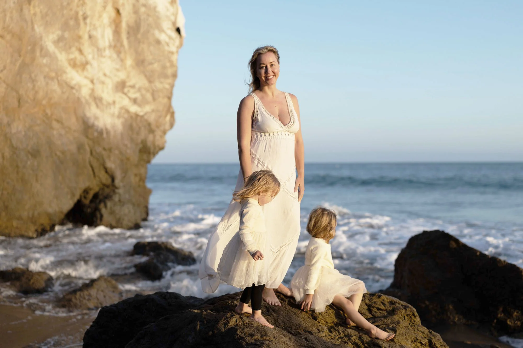 A woman with three children on a rocky beach near the ocean, with a large rock formation on the left and blue sky above.