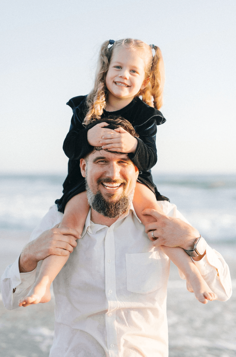 A man with a beard carrying a young girl on his shoulders at the beach, both smiling and enjoying the moment during sunset.
