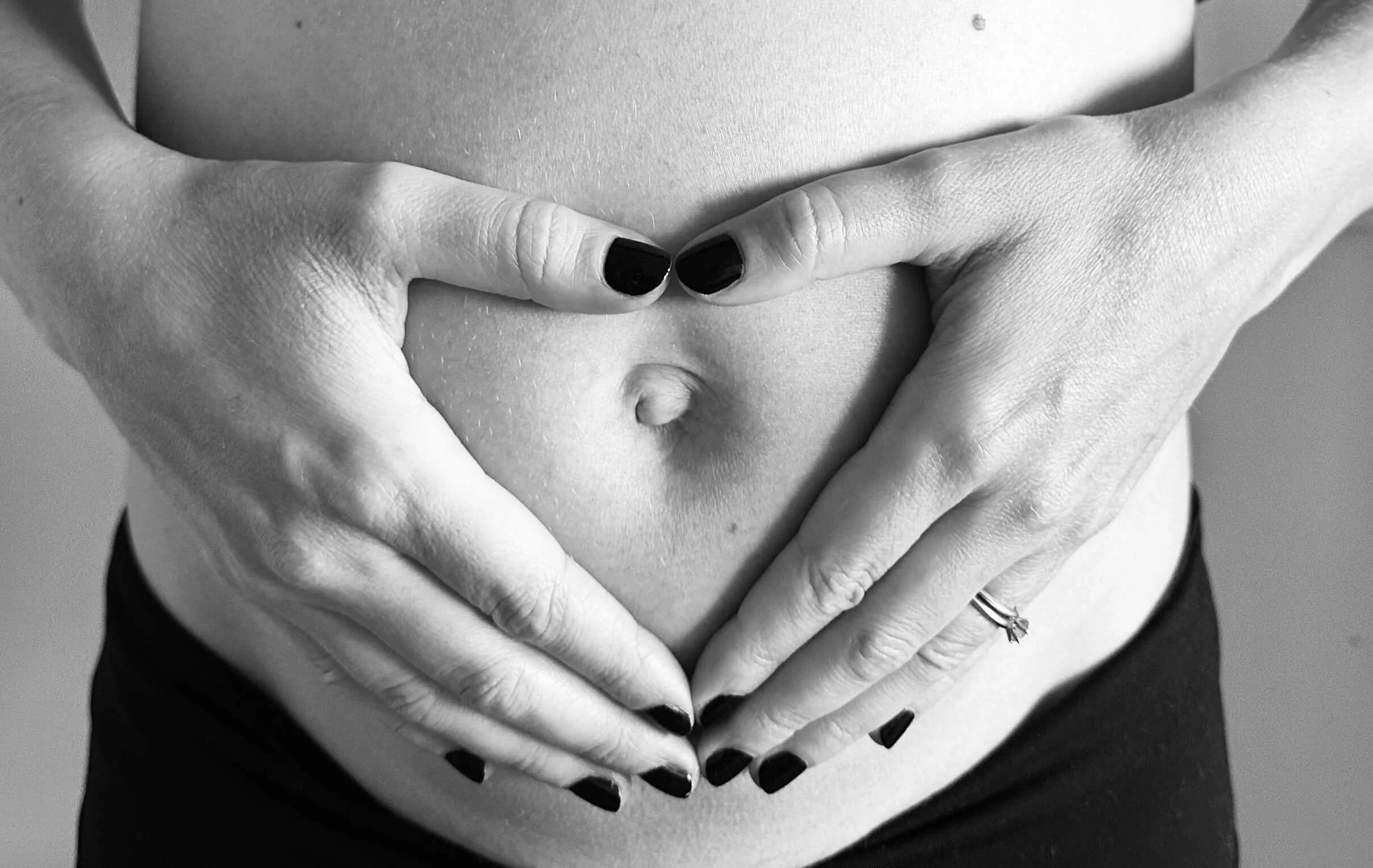 Black-and-white photo of a person with black nail polish making a heart shape with their hands on their bare stomach, with a ring on one finger.