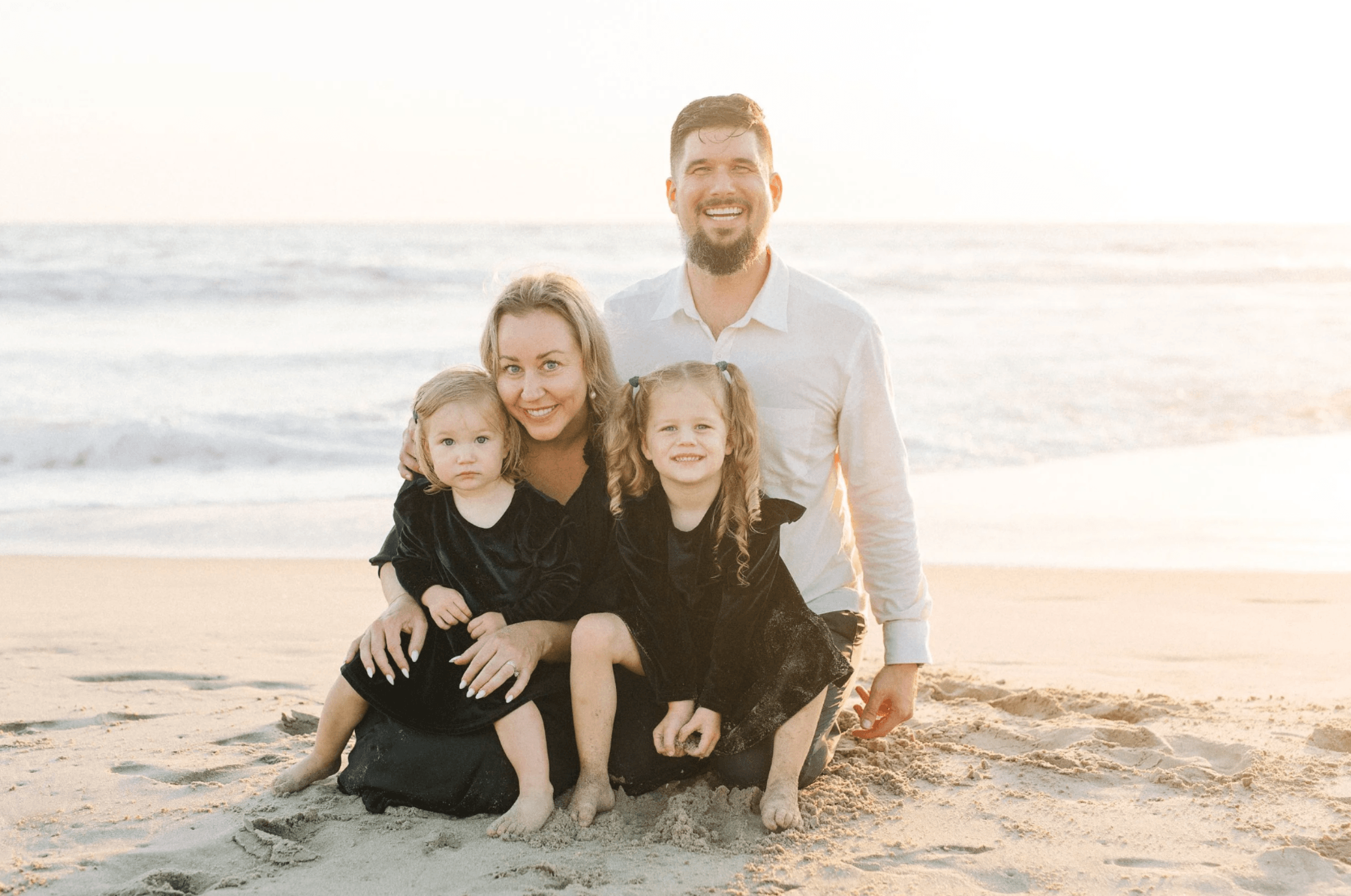 Family of four on a beach during sunset, with a man, woman, and two young girls smiling and sitting on the sand.