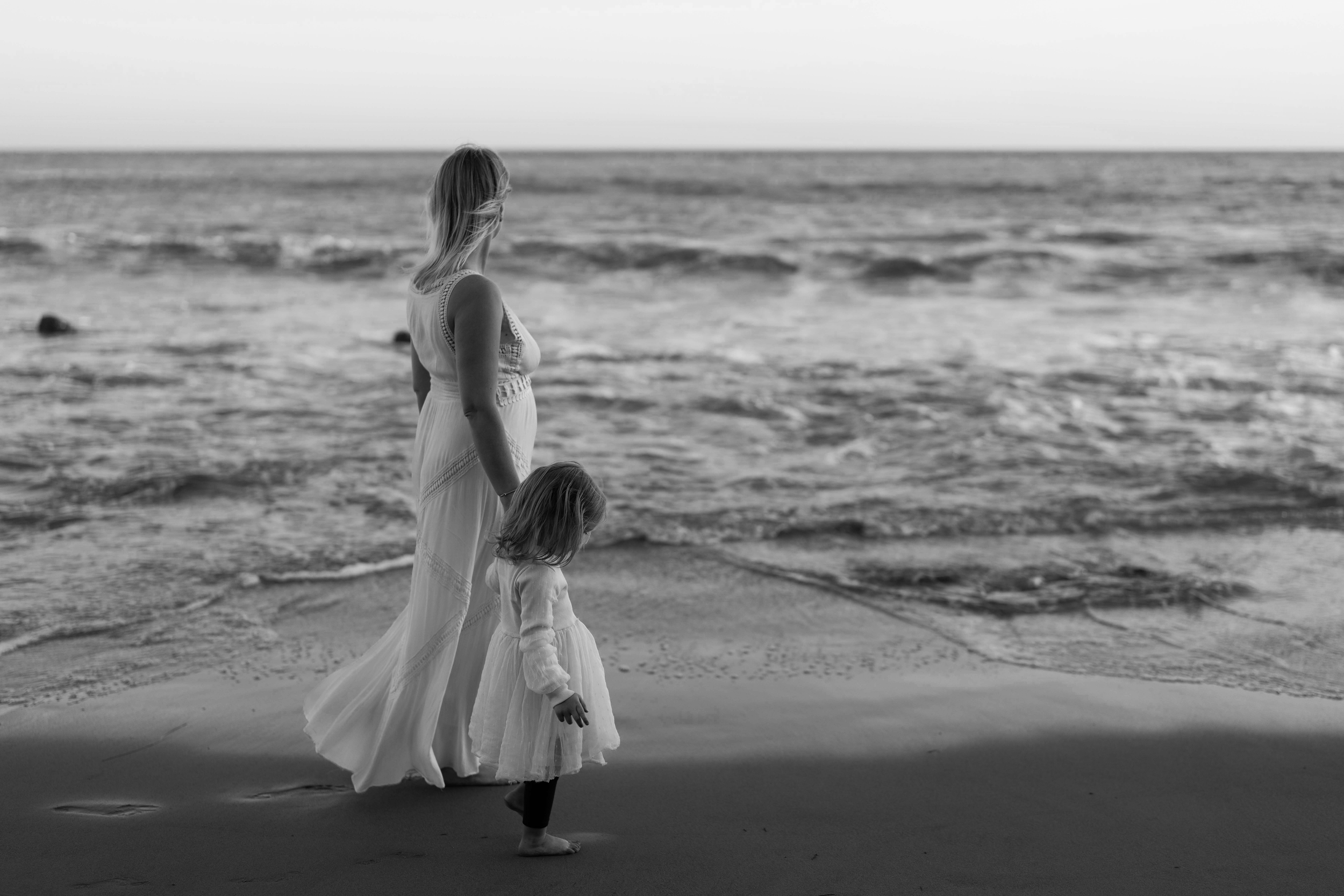 A woman and two young girls walking along the beach. The woman is holding one girl’s hand, and the other girl walks ahead, all dressed in flowing white dresses. The scene is in black and white, with waves in the ocean in the background.