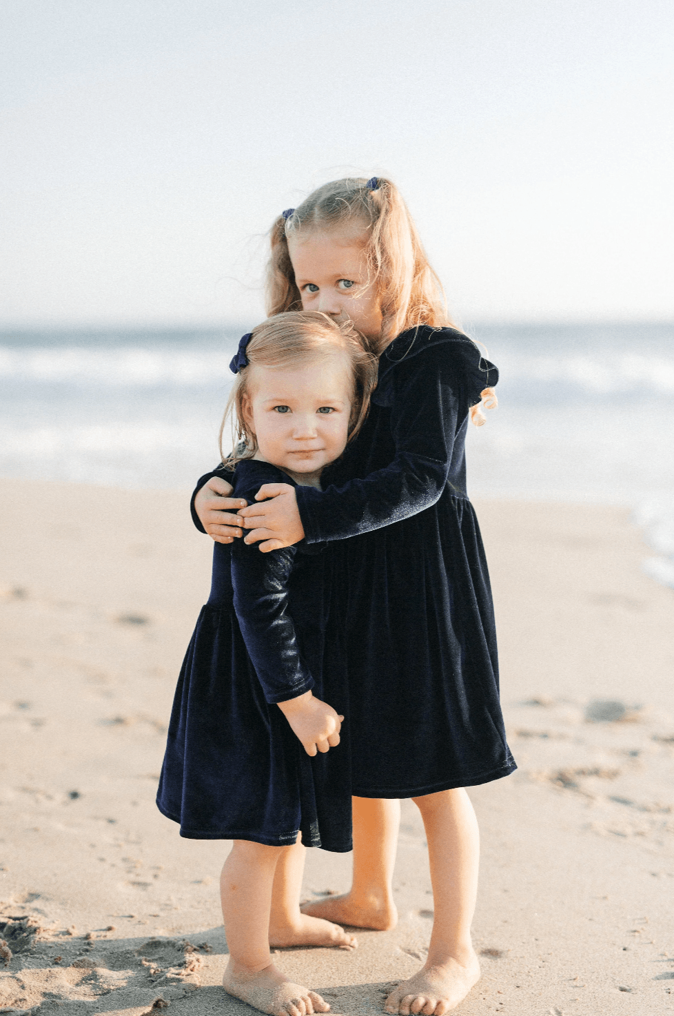 Two young girls in matching navy velvet dresses hugging each other on a sandy beach with ocean waves in the background.