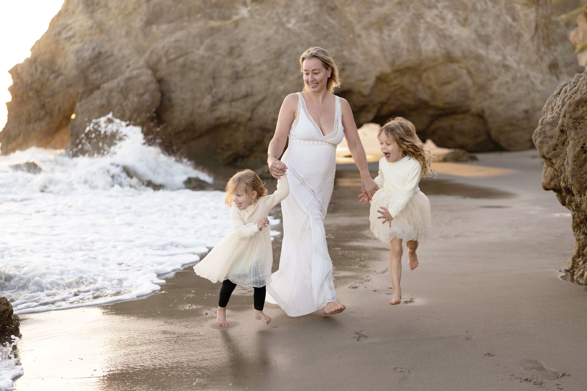 A woman and three young girls walking along the shoreline of a beach with rocky formations and an arch in the background, all dressed in light-colored clothing.