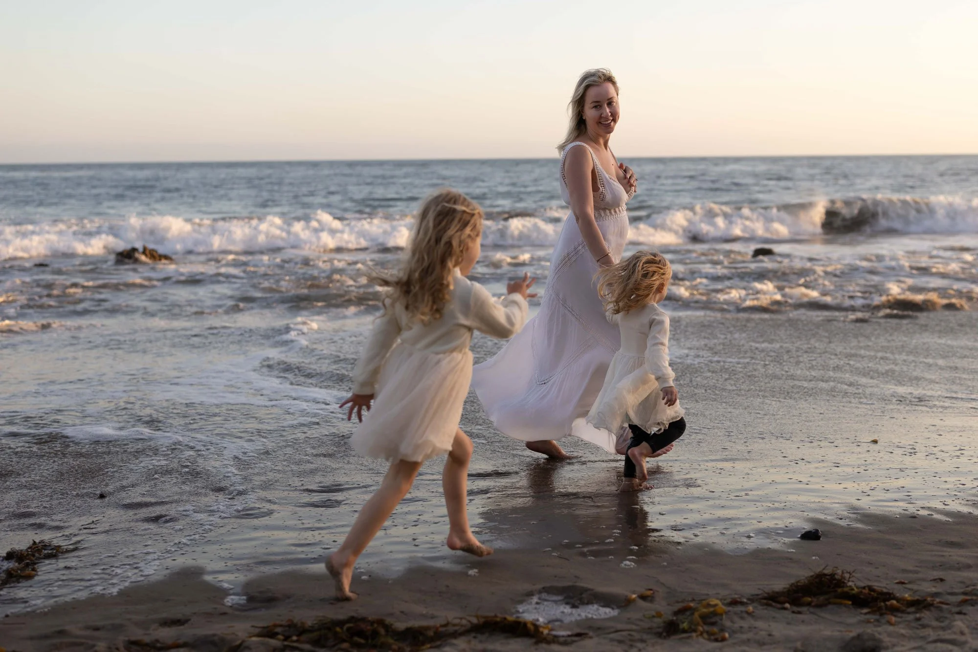 A woman in a white dress and two young girls in light-colored dresses running and playing on a beach near the ocean at sunset.