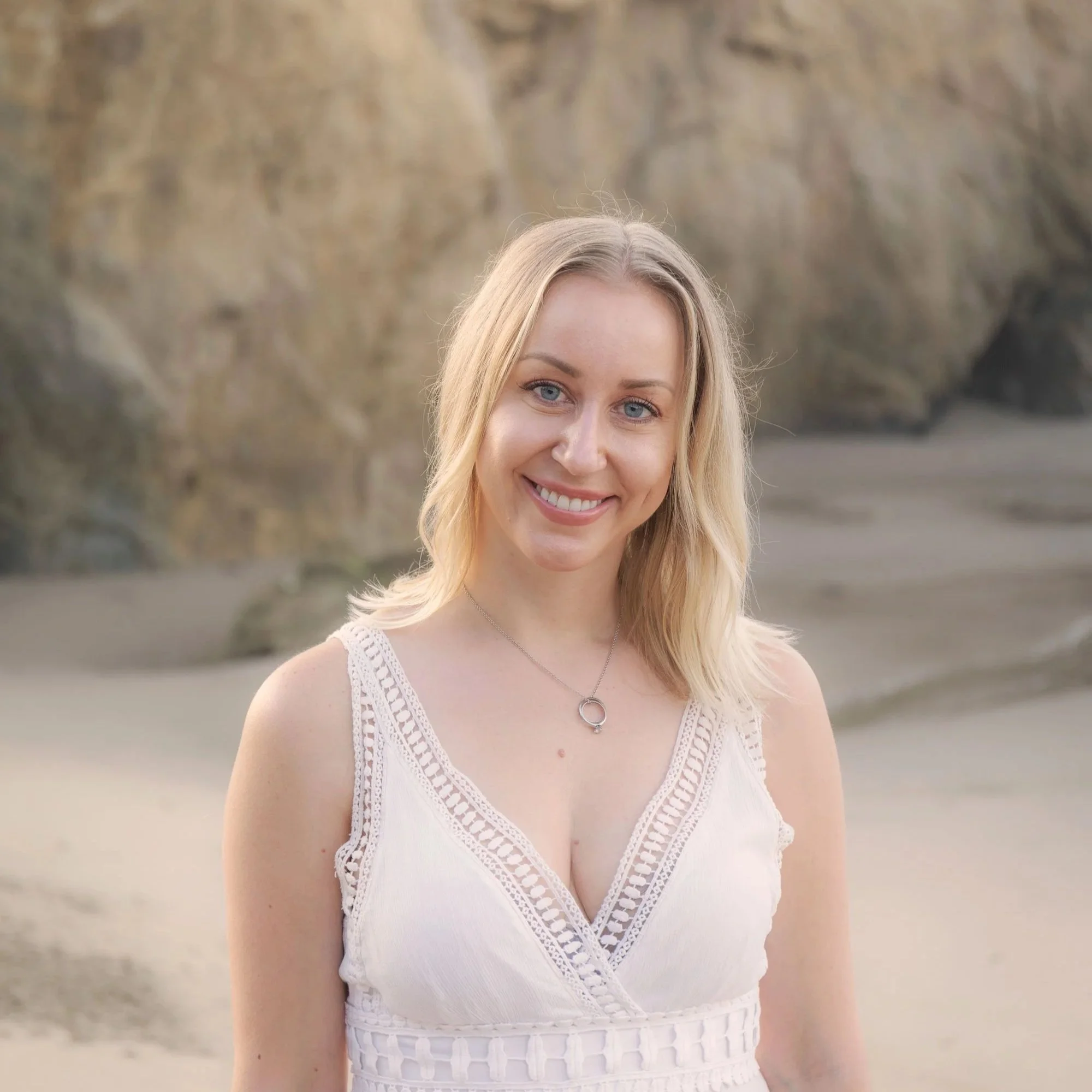 Close-up of a woman with blonde hair and blue eyes smiling, wearing a white sleeveless dress with lace details, outdoors with a rocky background.