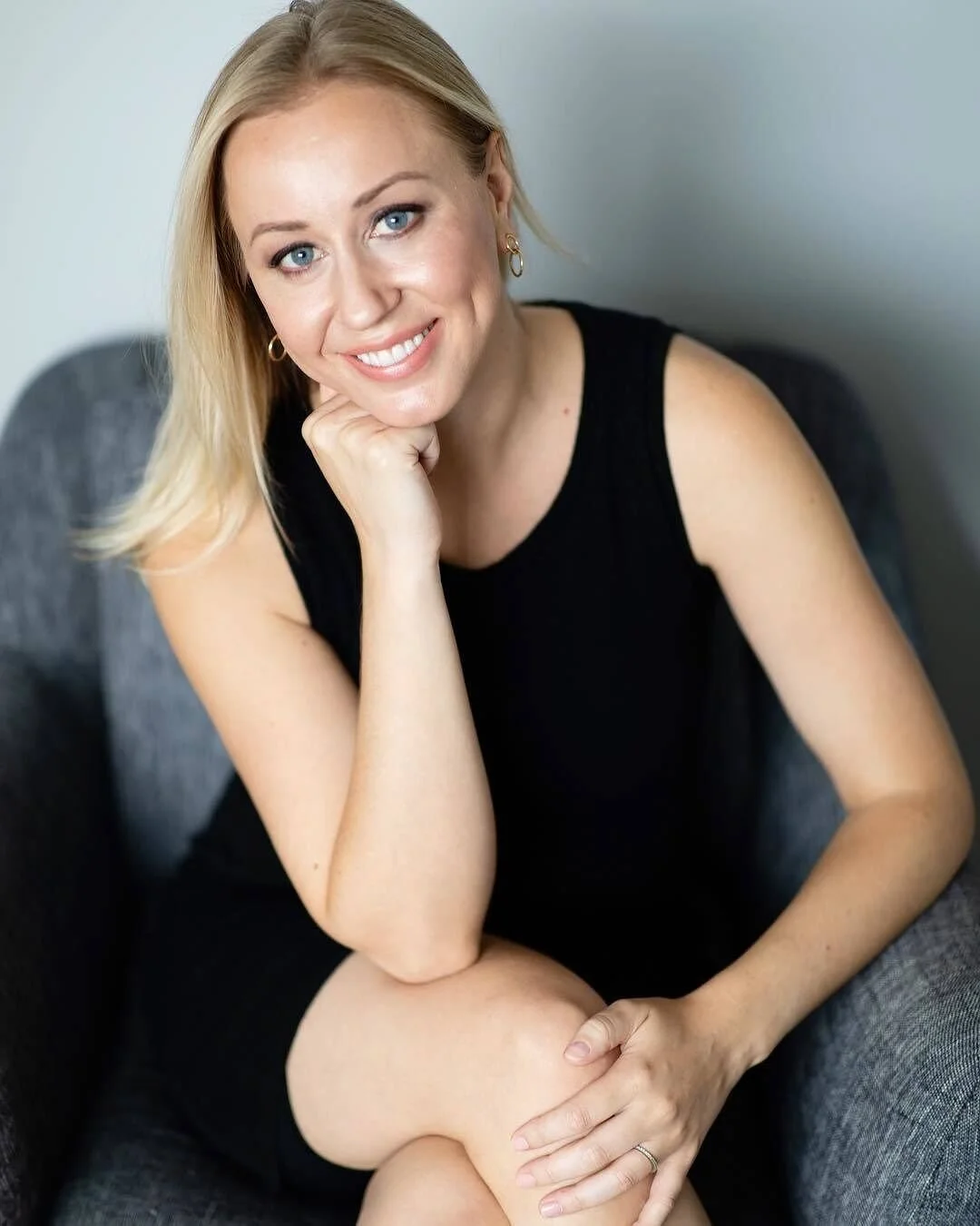 A woman with blonde hair wearing a black sleeveless top, sitting on a gray armchair, smiling with her chin resting on her hand, against a plain background.