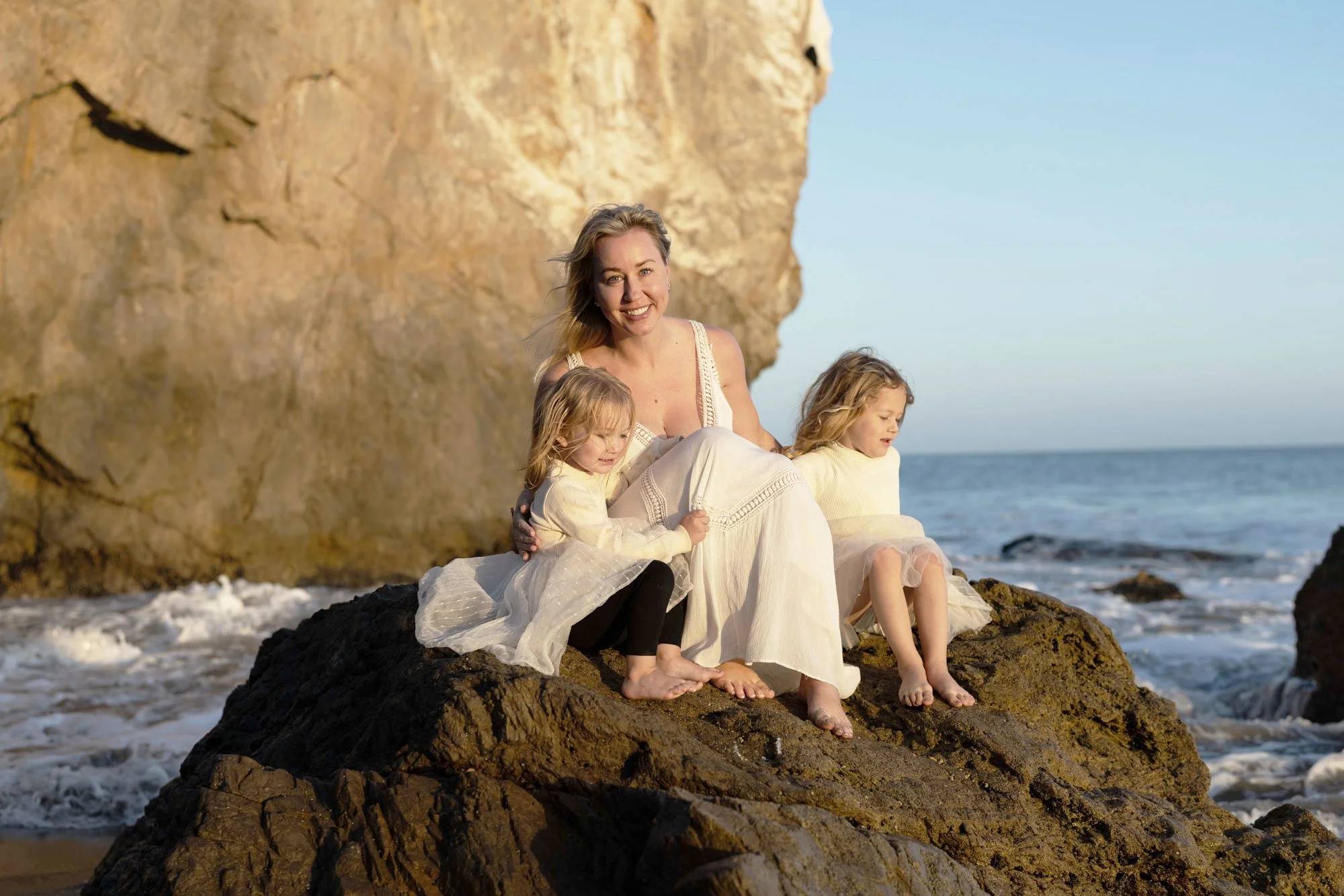 A woman with two young girls sitting on rocks at the beach near a large cliff, with waves crashing at their feet and the ocean in the background.
