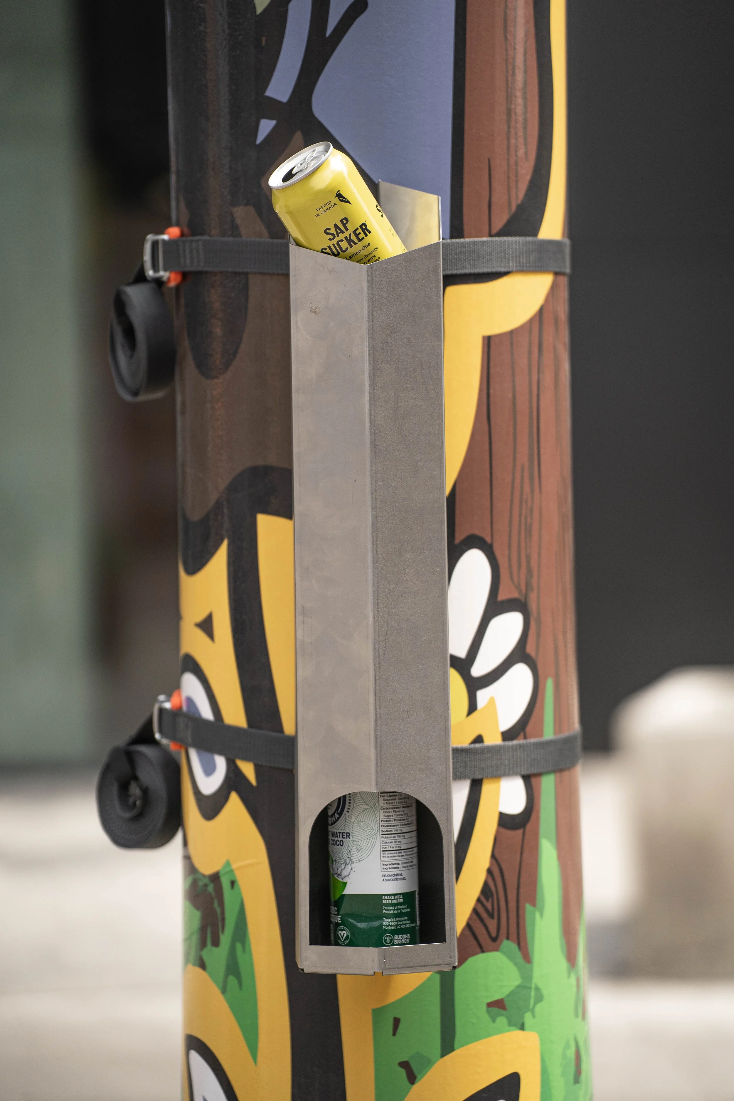 An image of the Can Companion protyope attached to a pole. It is a cylindrical shape made out of metal and there are cans inside it.