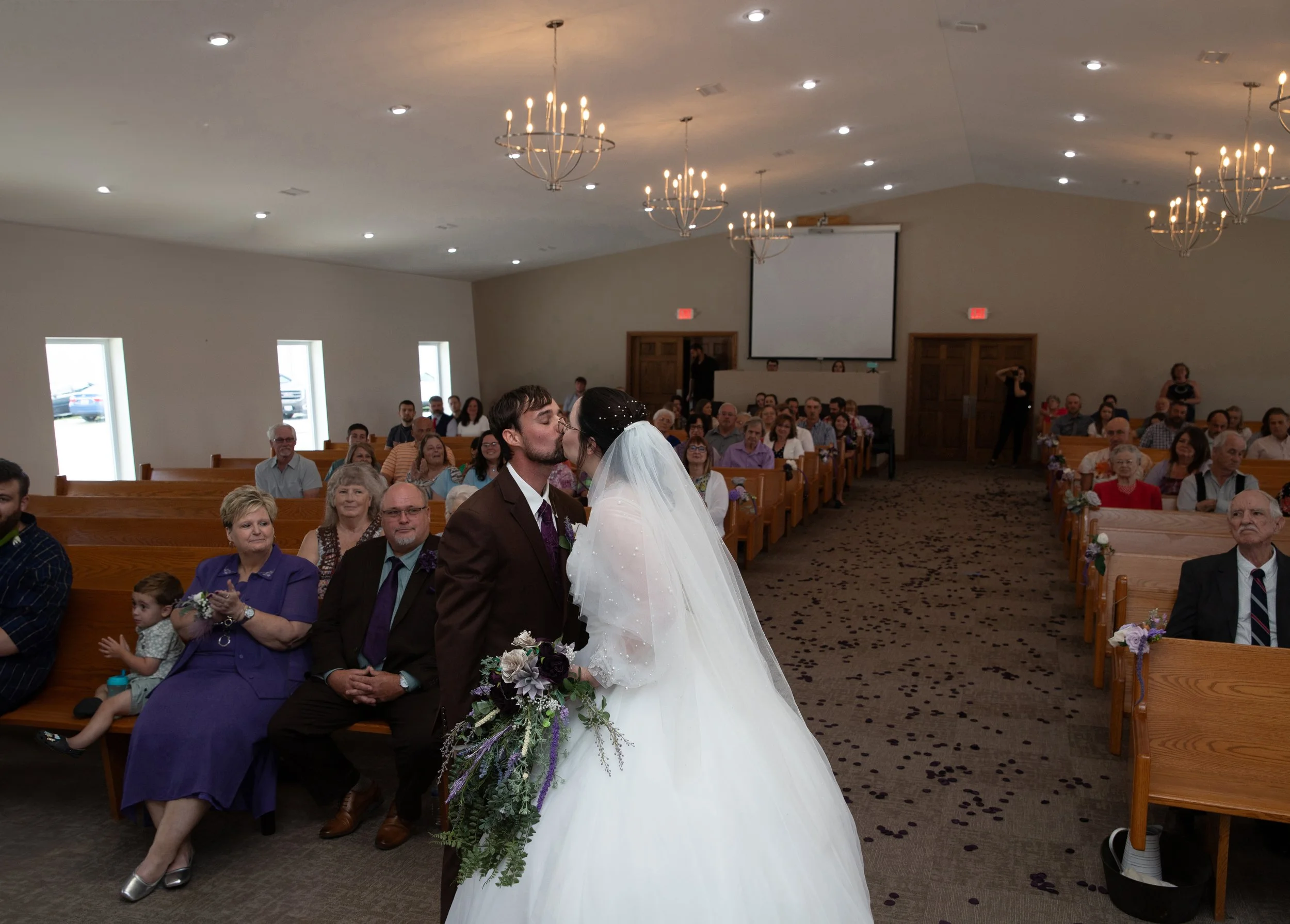 Removed the officiant from the kissing image by seamlessly blending pieces of the background from another photo and skillfully recreating the windows on the wall for a natural, cohesive result.