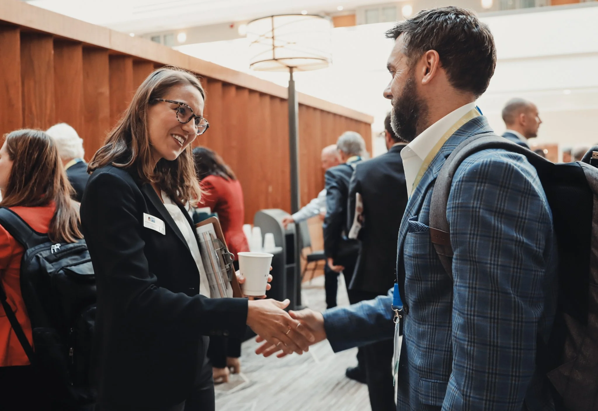 A woman in glasses and a black blazer shaking hands with a man in a checkered blazer and white shirt at a professional event.