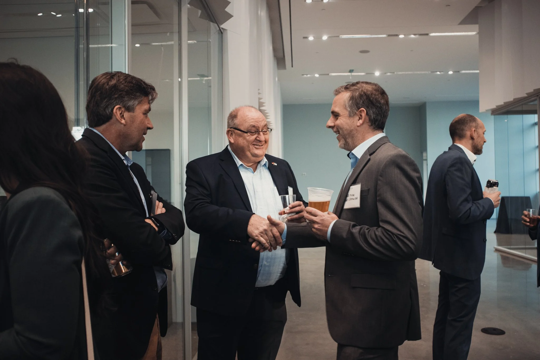 Six people in business attire are engaged in conversation at a networking event. Two men are shaking hands while holding drinks. The background shows a modern office or conference center.