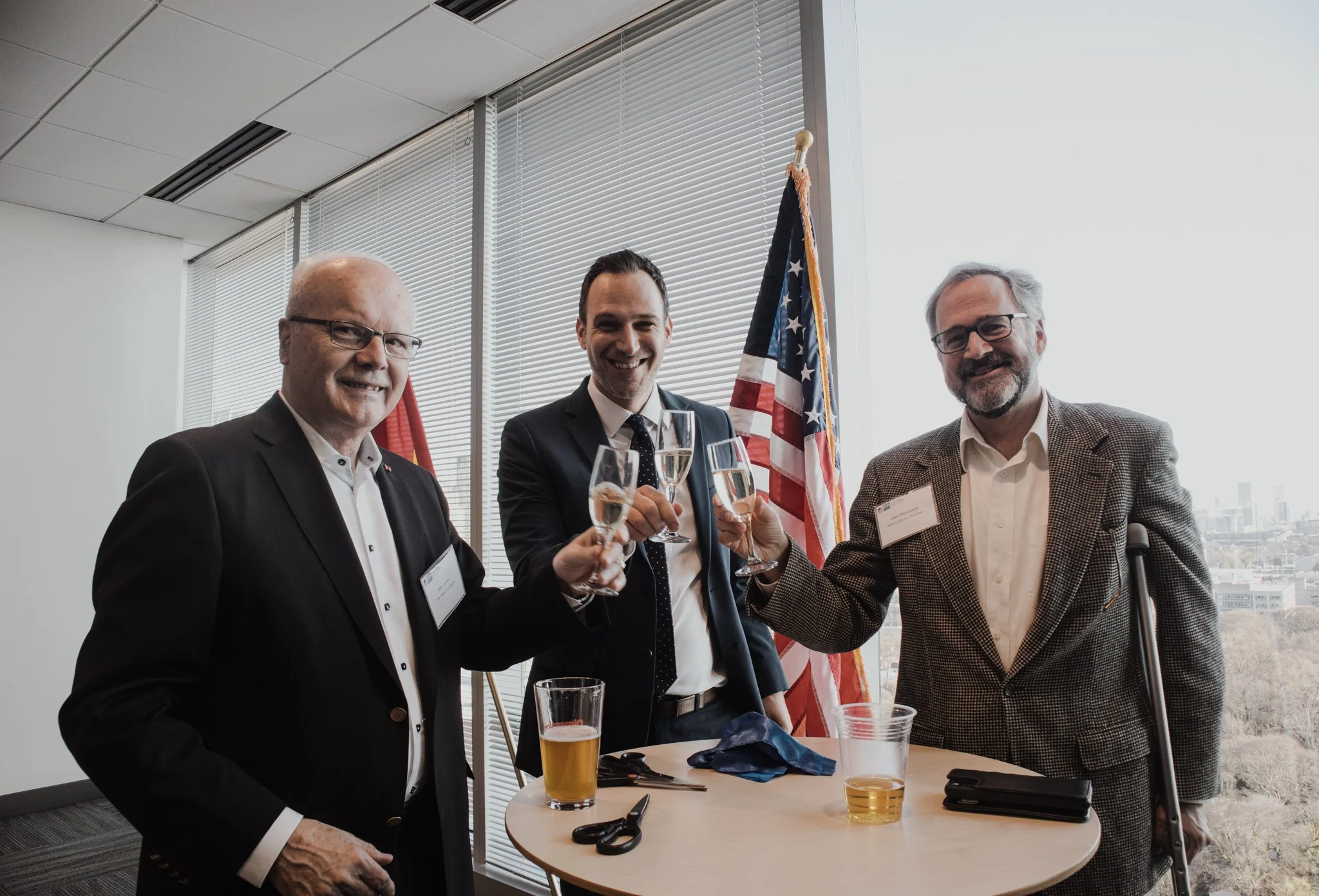 Three men in business attire are celebrating with drinks in an office setting. One man with glasses is smiling, the second man in a suit is raising a glass of champagne, and the third man with gray hair and a beard is smiling. There are American flag